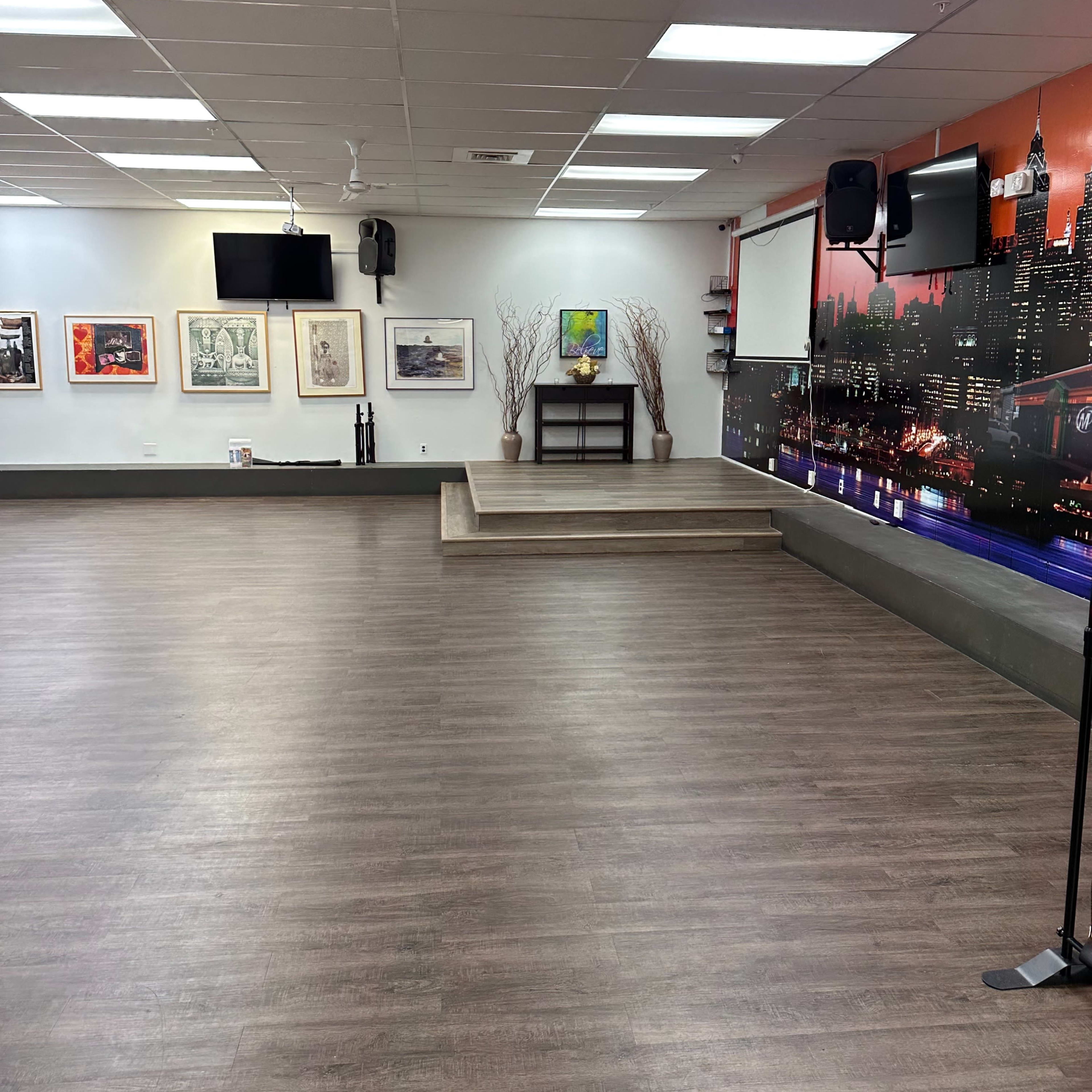 A spacious indoor room with a wooden floor, a small elevated stage at one end, wall-mounted televisions, and artwork hanging on the walls.