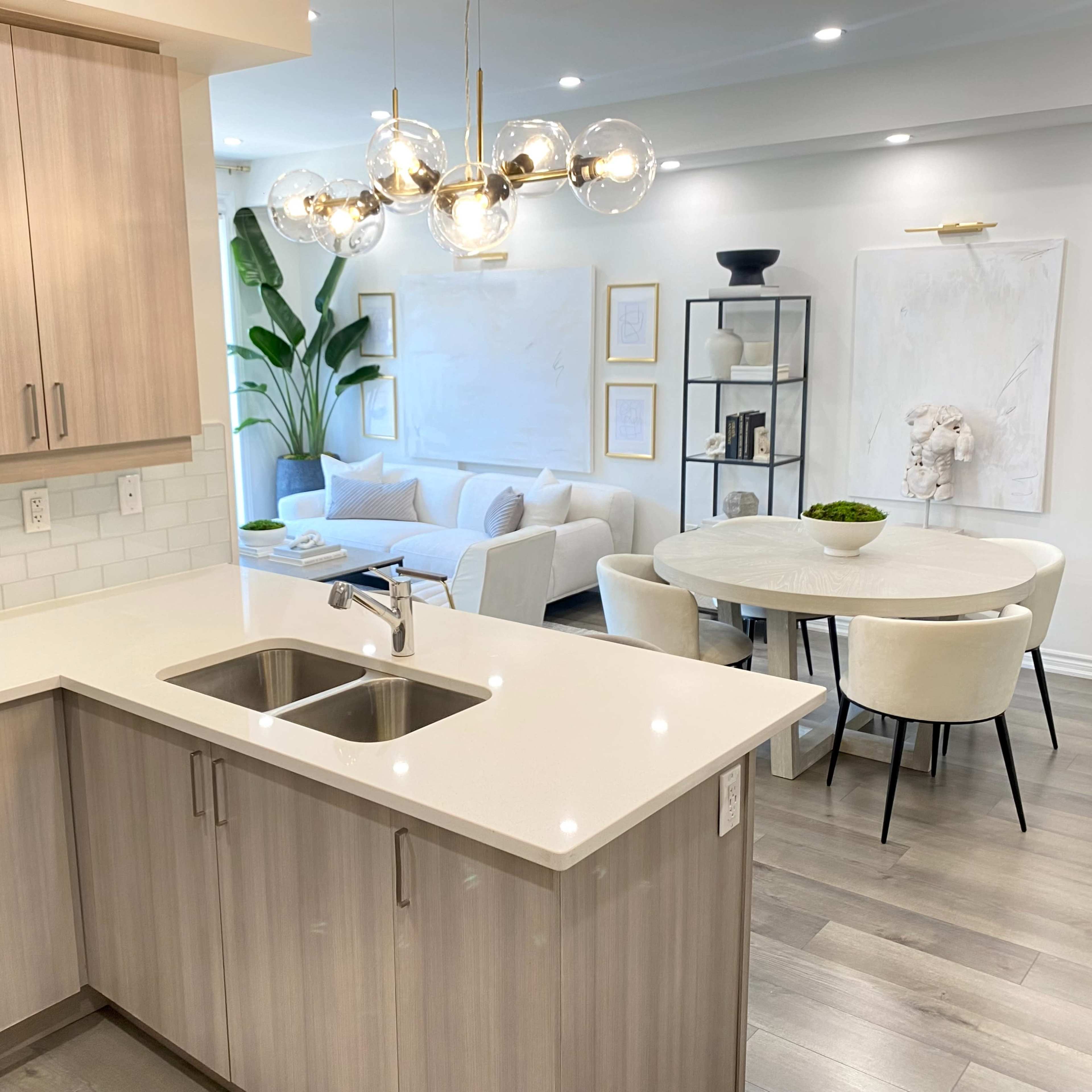 A modern kitchen with a double sink and light wood cabinetry opens up to a stylish living area featuring a round dining table and a comfortable seating arrangement.