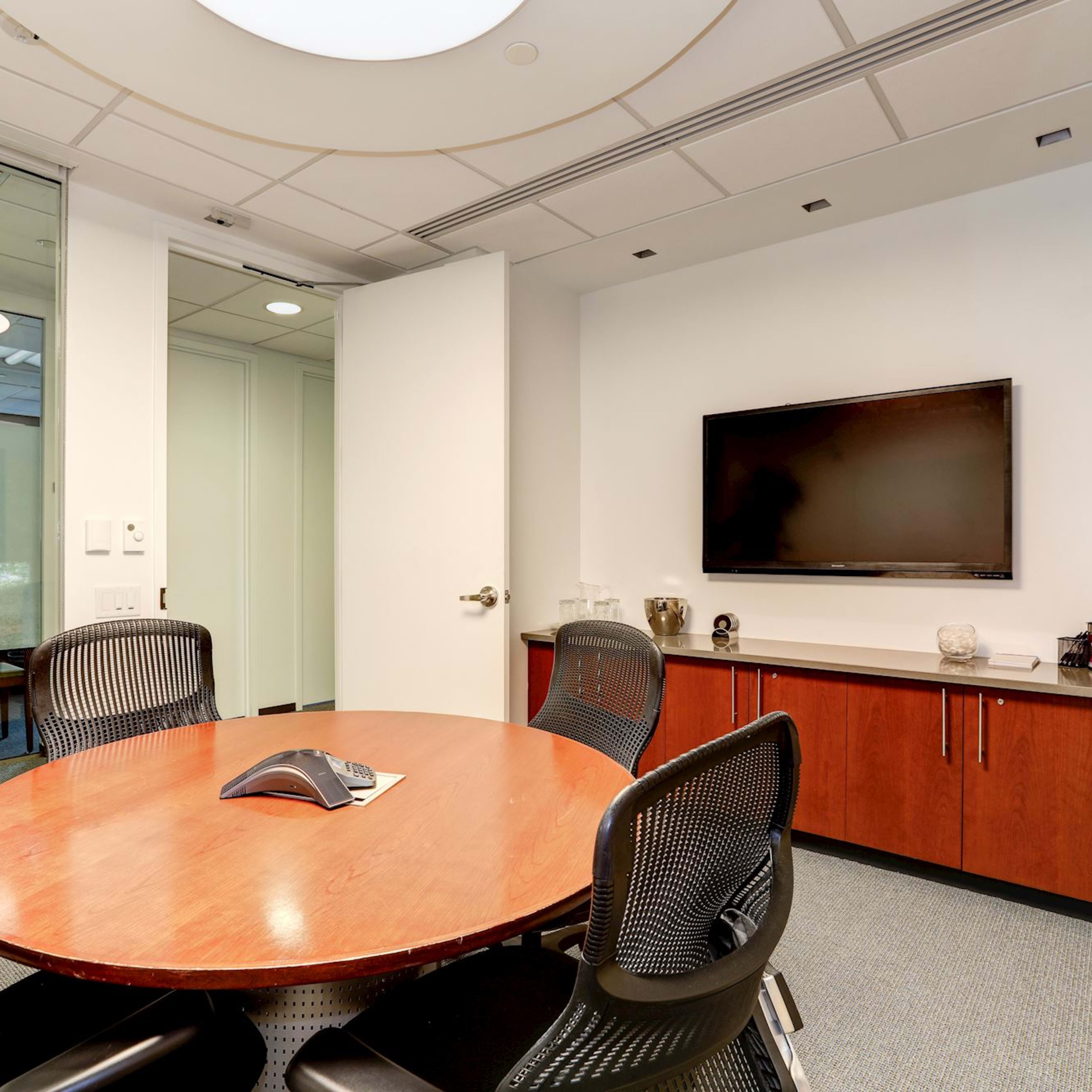 A modern conference room with a round wooden table surrounded by black mesh chairs, a television mounted on the wall, and glass partitions leading to another area.