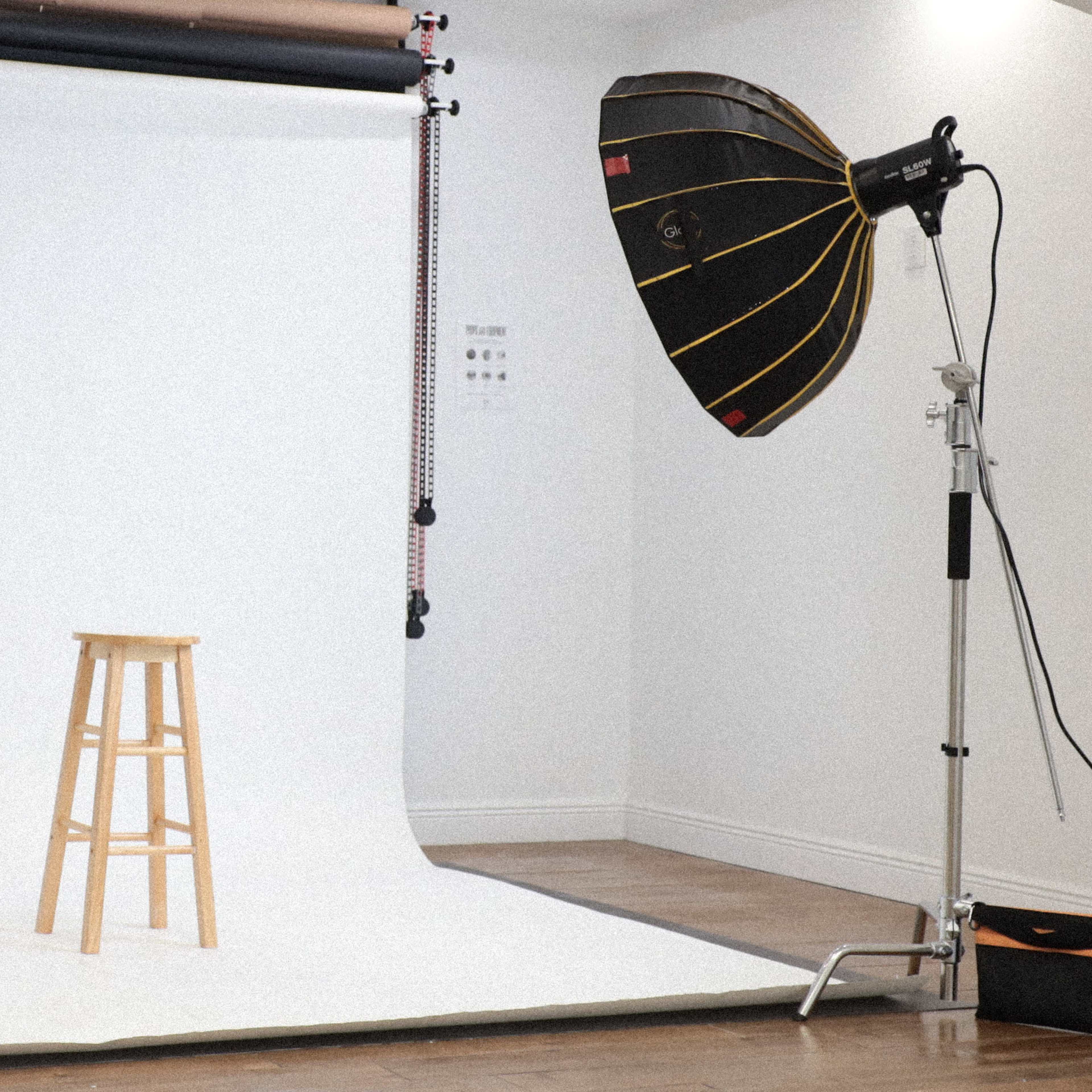 A photography studio setup featuring a white backdrop, a wooden stool in the center, and two large lighting umbrellas positioned on either side.