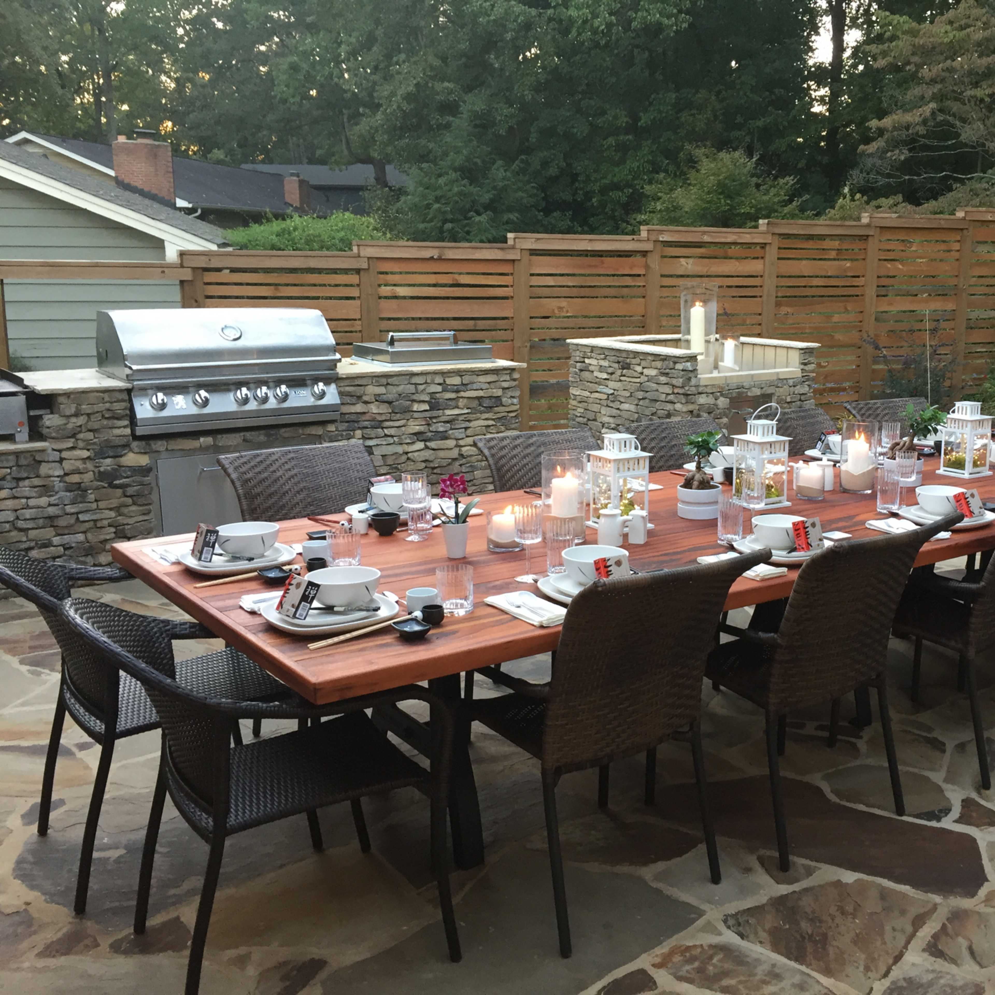 A long dining table is set outdoors with dishes and lanterns, surrounded by a stone patio and greenery.