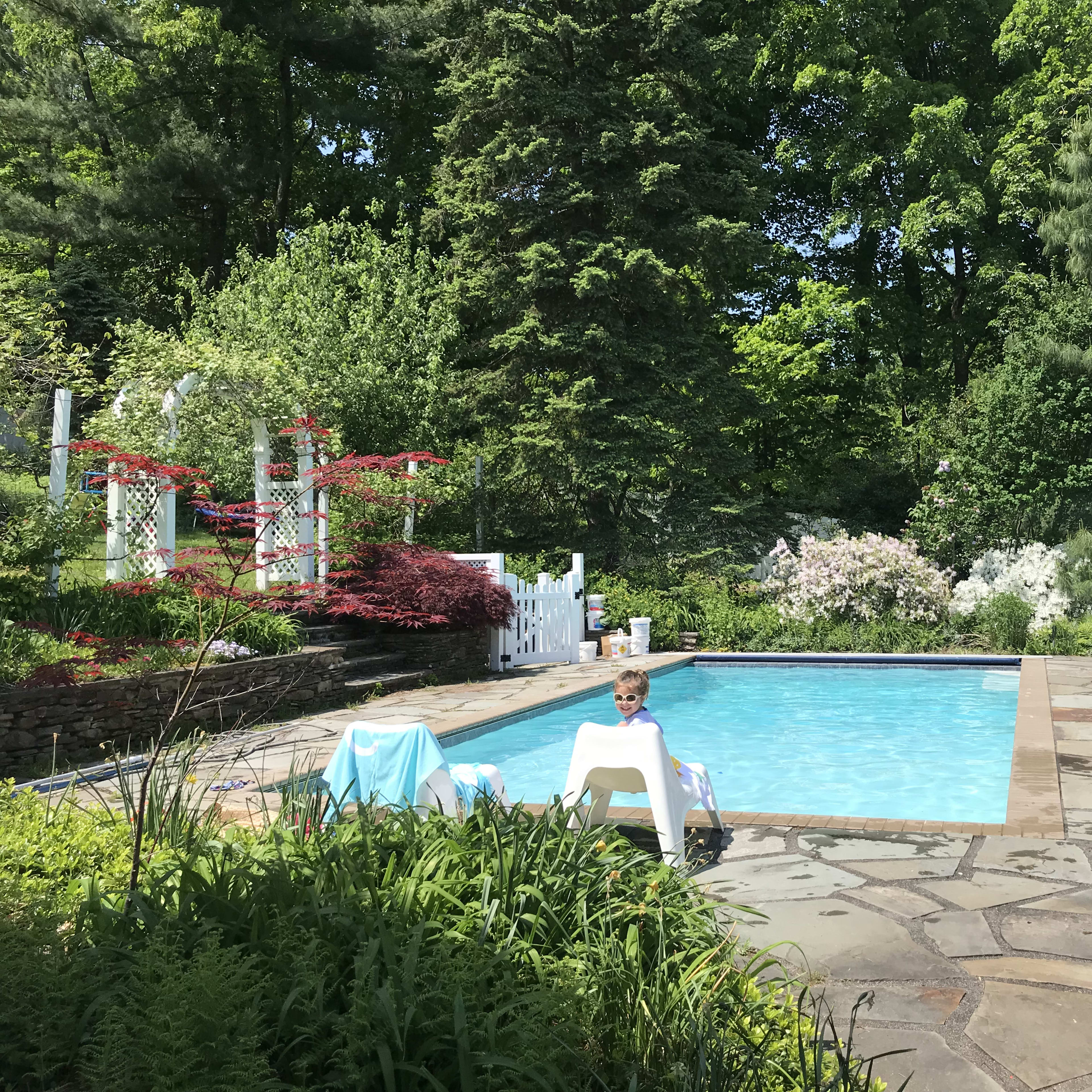 A child sits on a white lounge chair beside a swimming pool surrounded by greenery and colorful flowers.