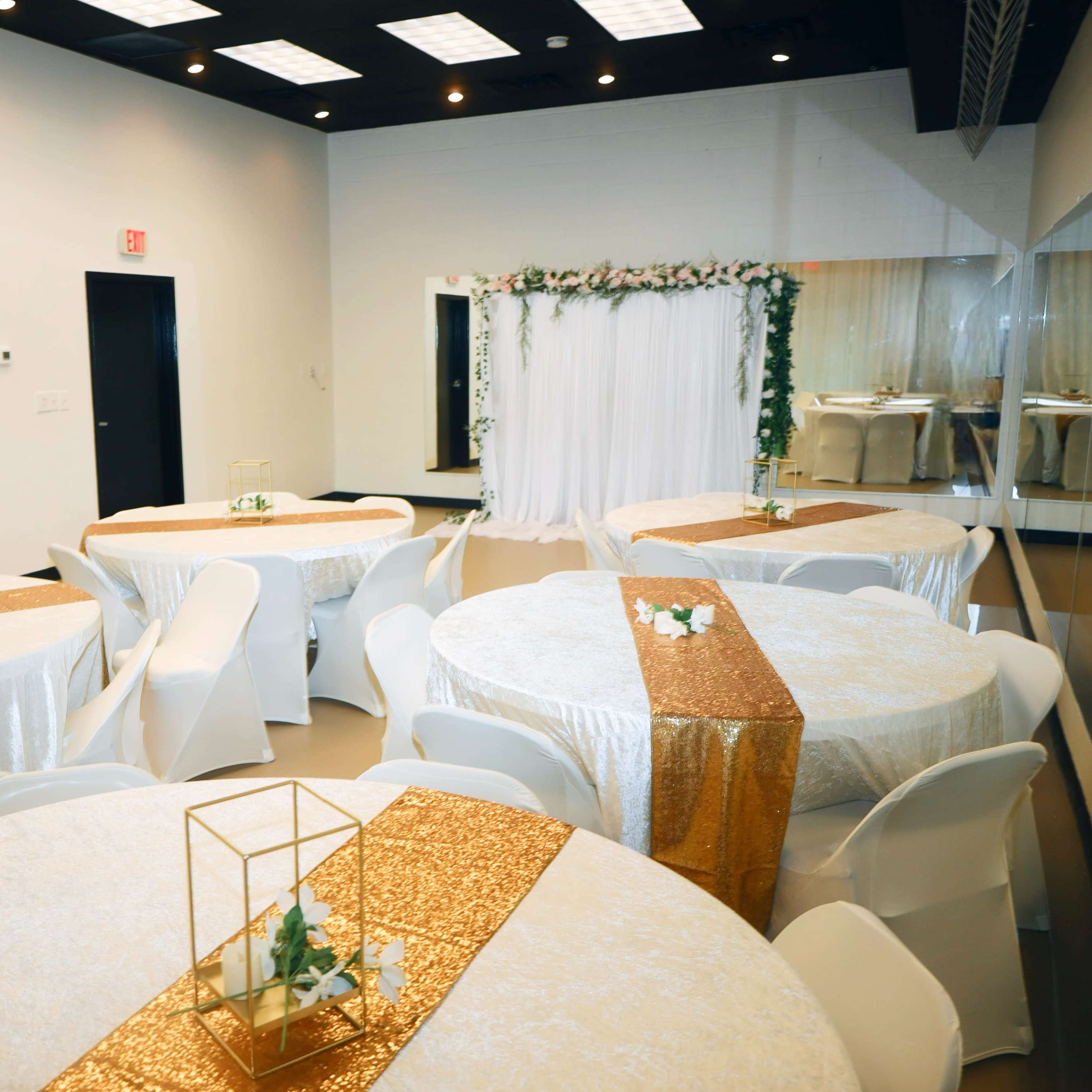 A decorated event space with several round tables covered in white tablecloths, gold runners, and a backdrop with greenery and drapery at one end.