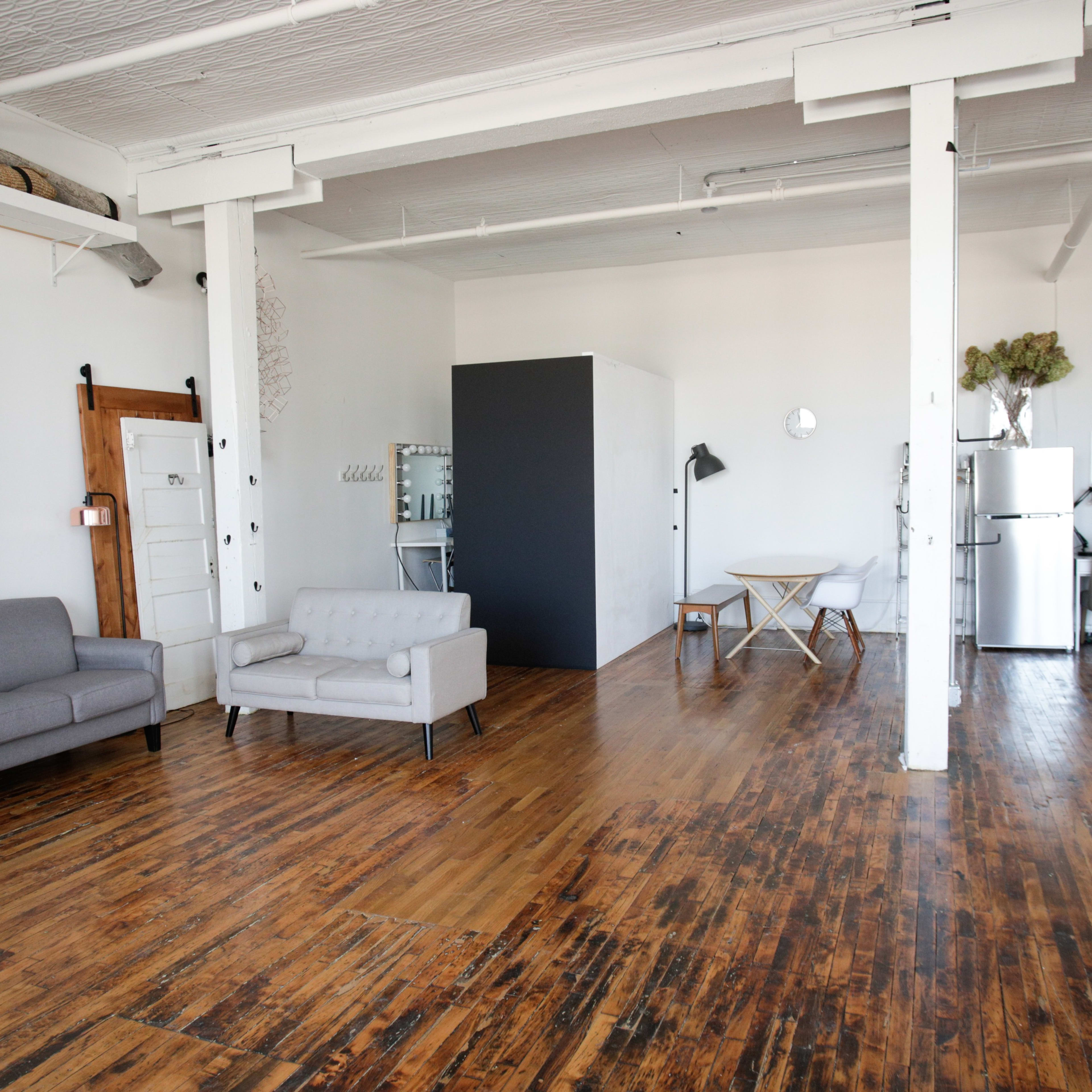 A spacious, minimalist room with wooden flooring, featuring a gray sofa, a small table, and various pieces of furniture against white walls.