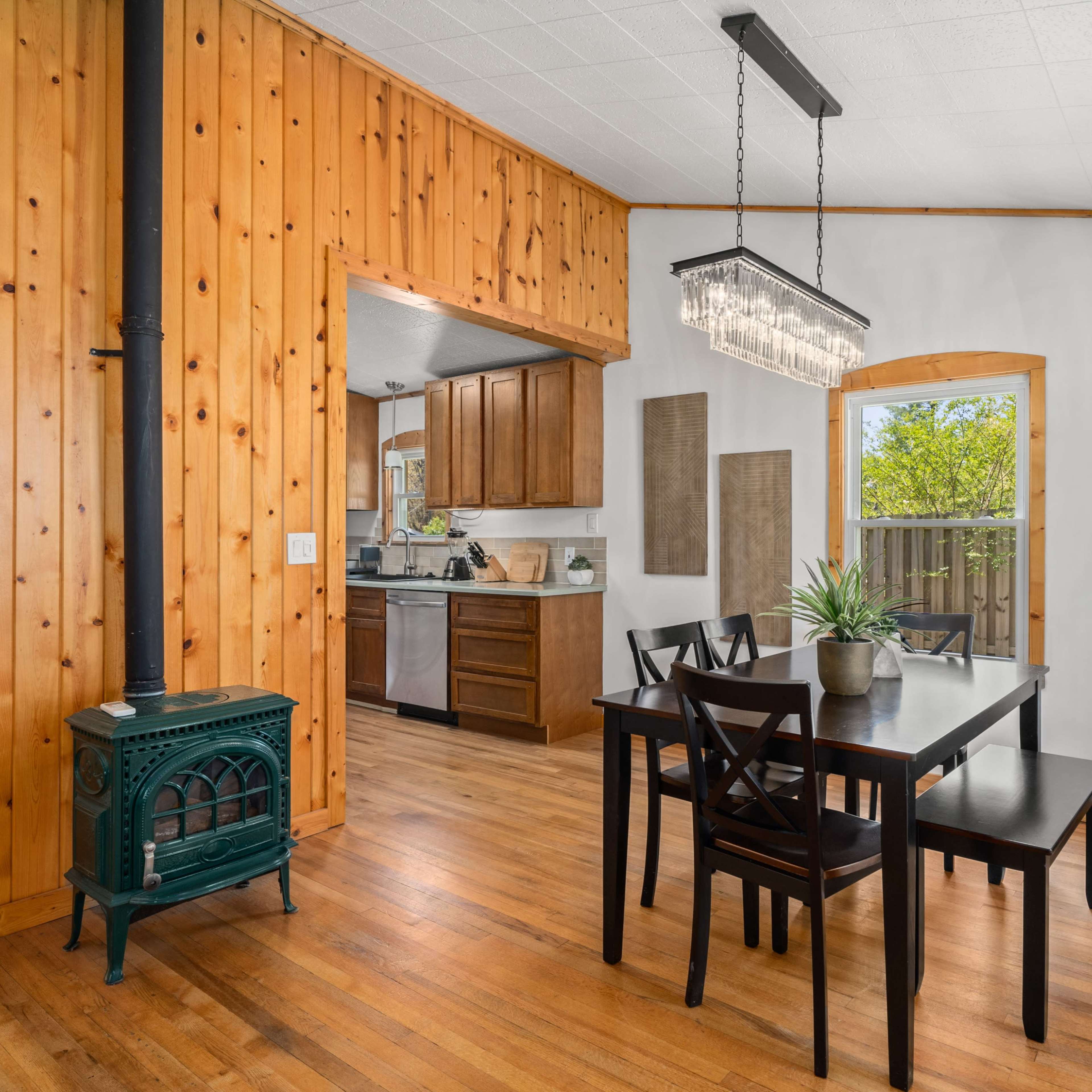 A dining area with a wooden table surrounded by black chairs, a green wood stove, and an adjoining kitchen area with wooden cabinets.