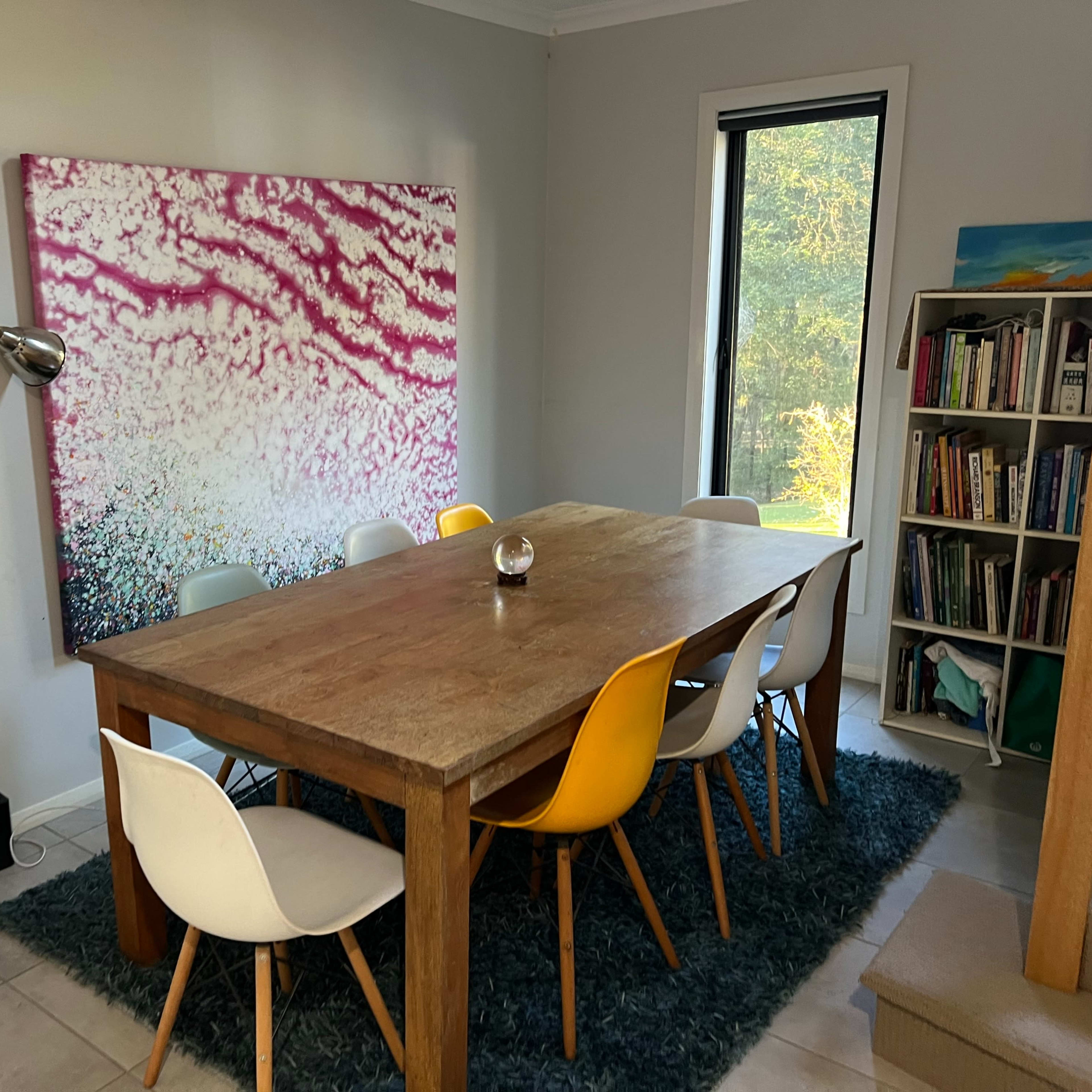A dining area features a large wooden table surrounded by colorful chairs, with a lamp and a bookshelf in the background.