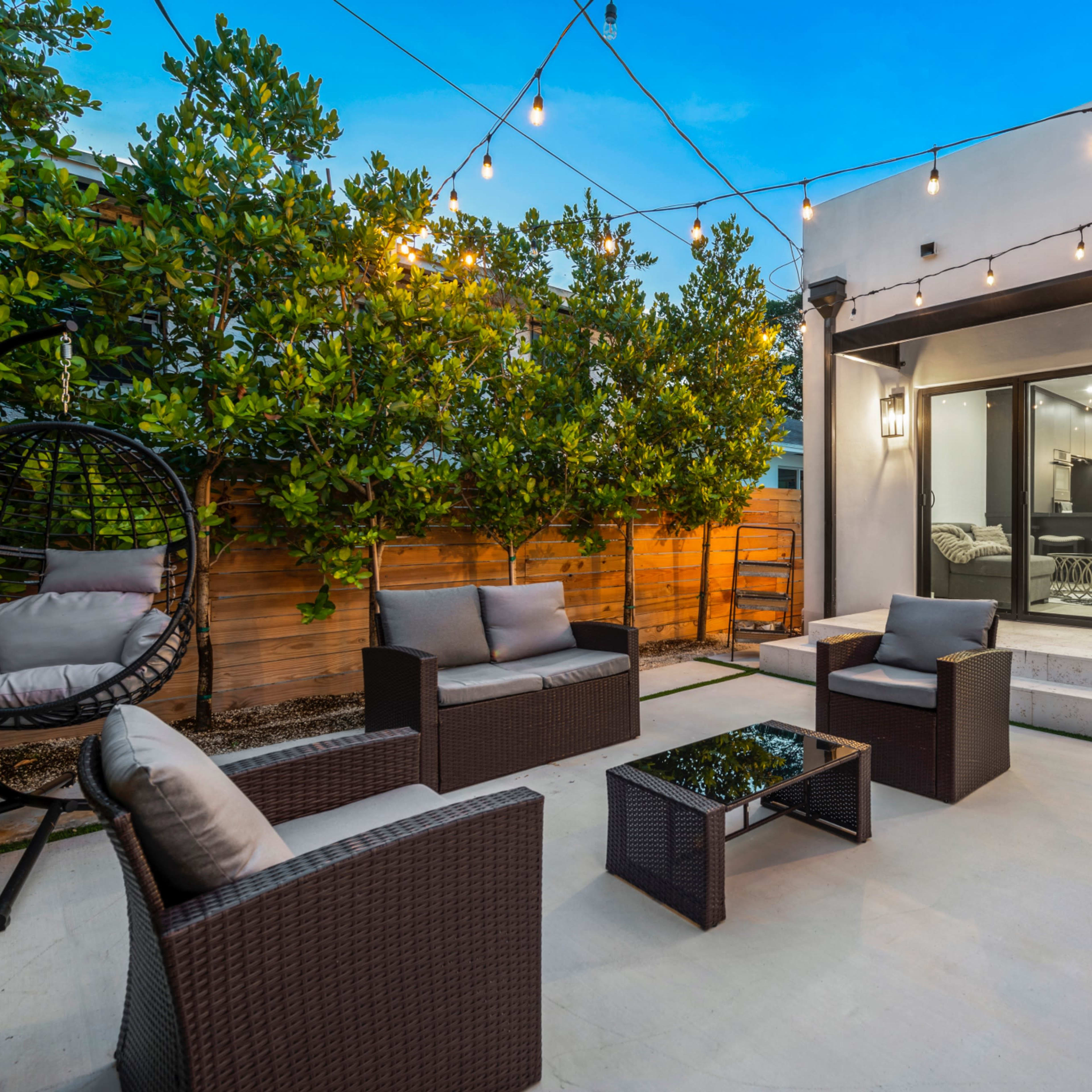A small outdoor seating area with four chairs and a glass table, surrounded by greenery and string lights.