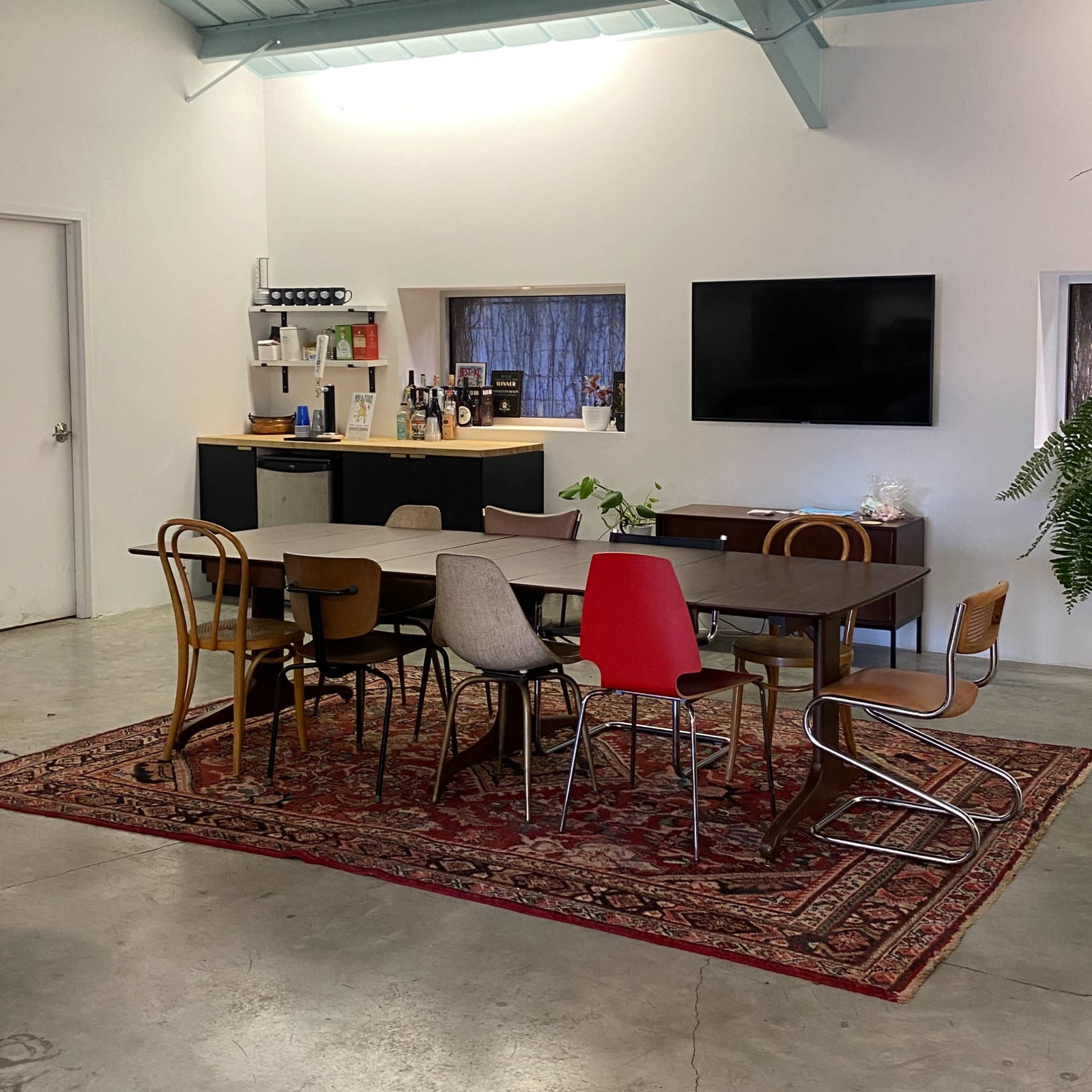 A spacious room with a table surrounded by various chairs, a bar area in the back, and a television mounted on the wall.