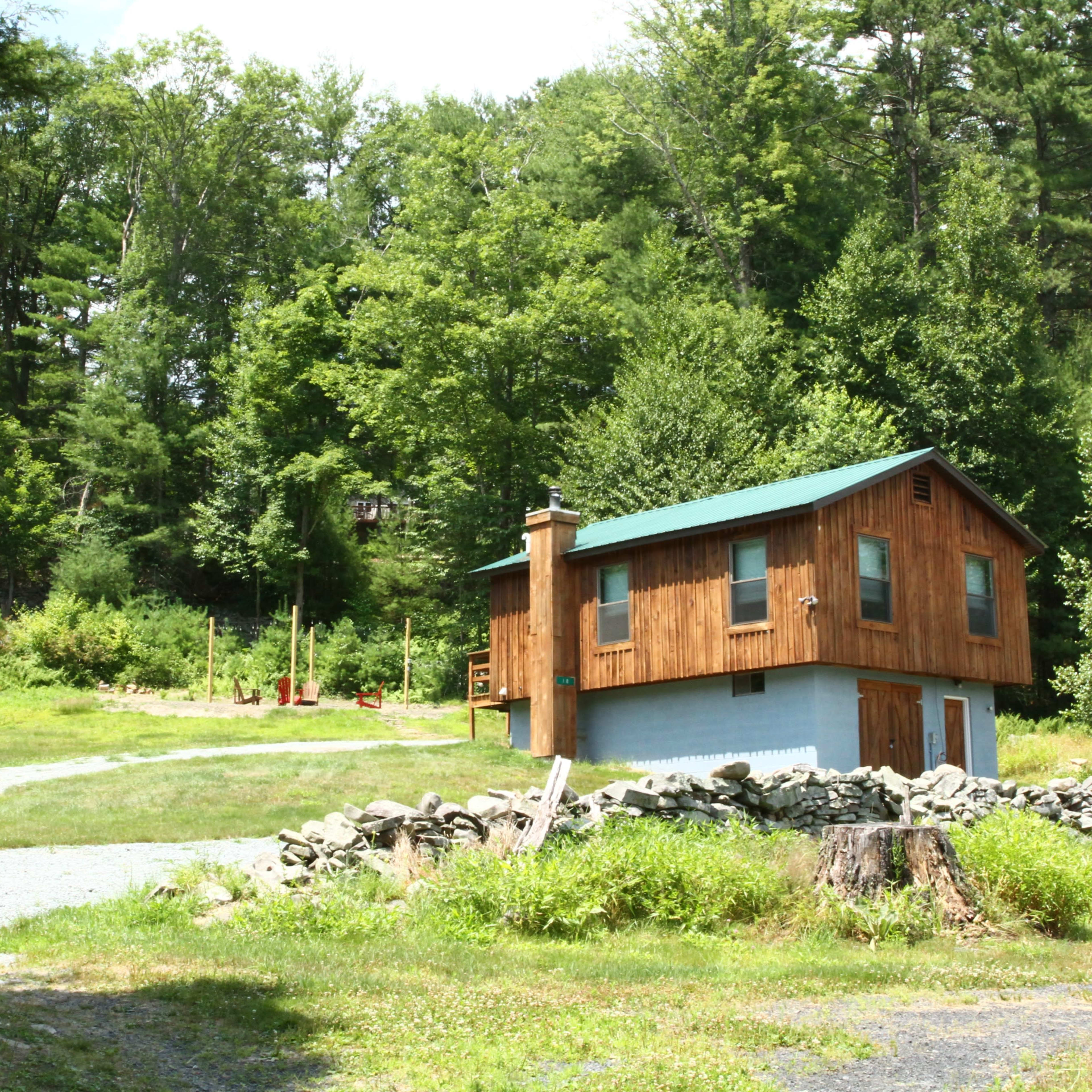 A wooden two-story cabin with a green roof is situated near a gravel path and surrounded by trees and shrubs.