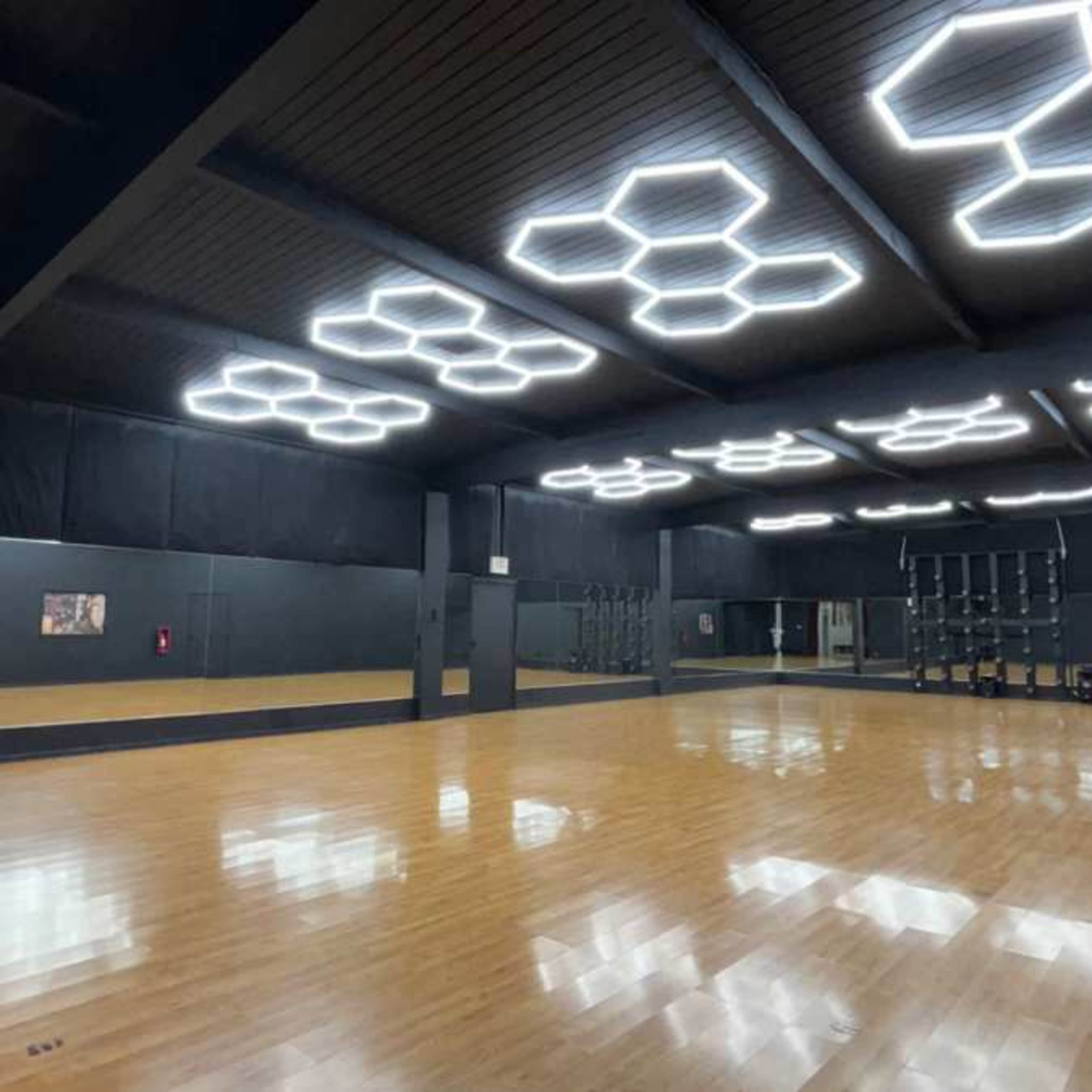 A spacious dance studio with a polished wooden floor, mirrored walls, and modern hexagonal ceiling lights.