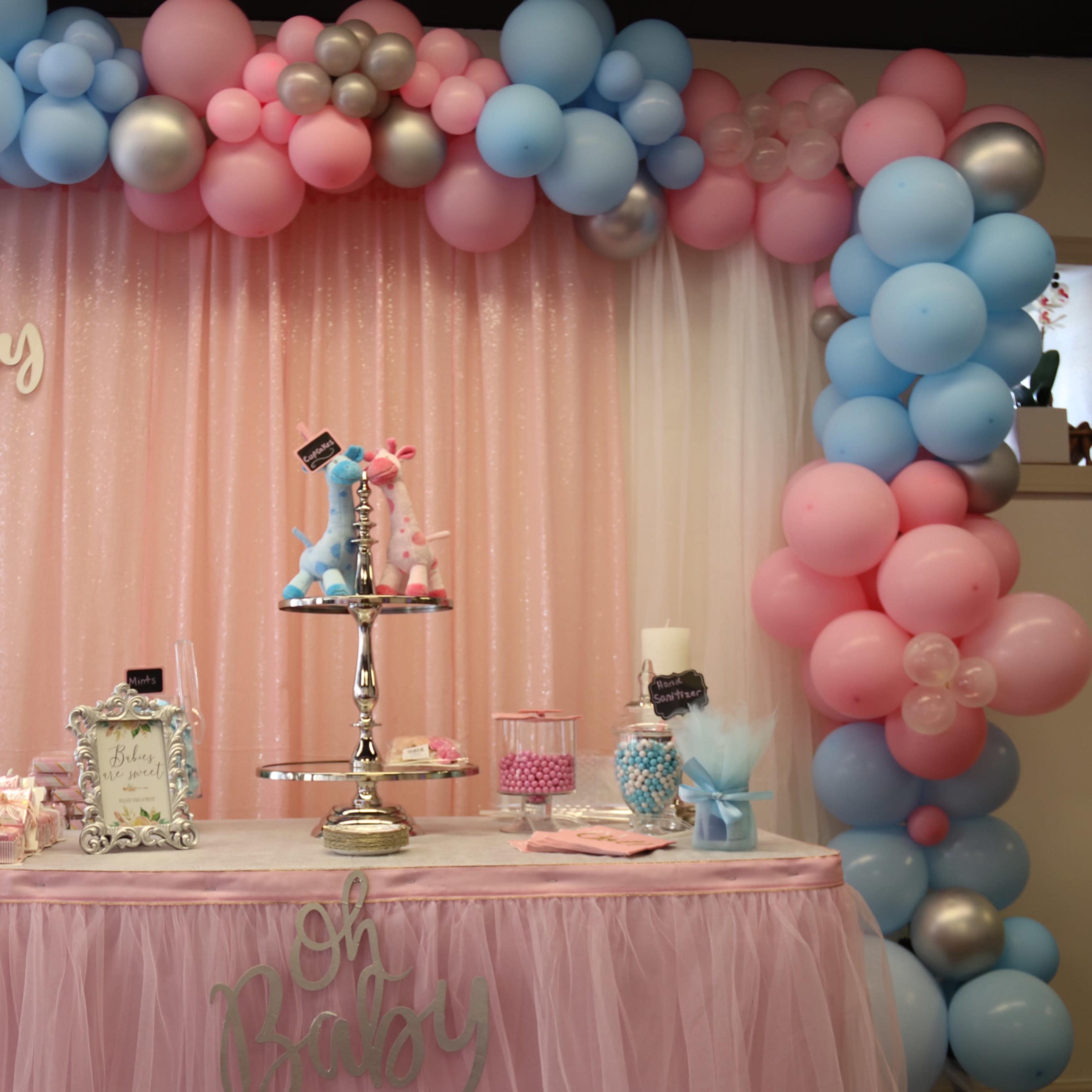 A decorated table with a pink tablecloth features a tiered stand of desserts, surrounded by a balloon arch in pastel colors and a sign reading "Oh Baby."