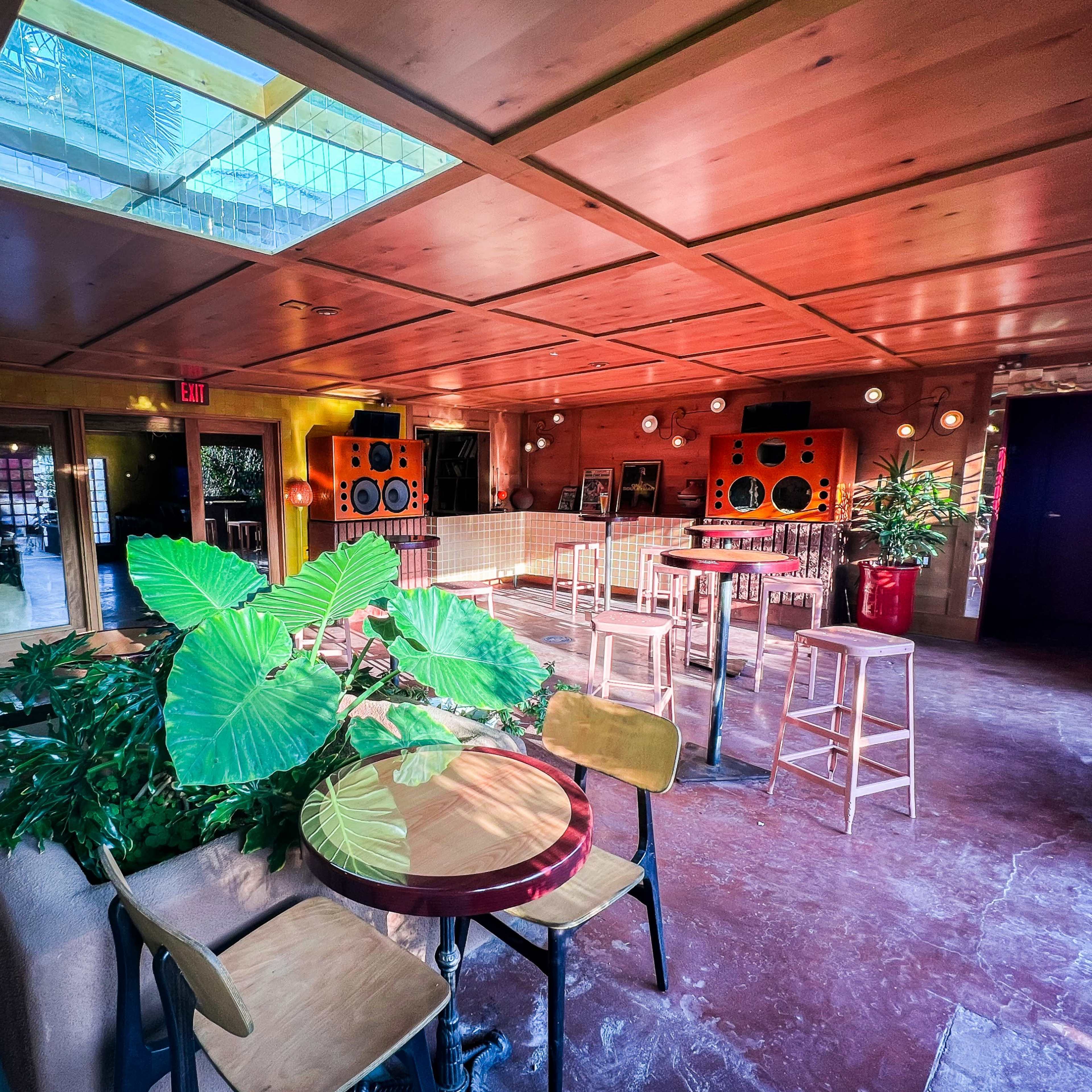 The interior of a colorful, spacious venue features wooden beams, green plants, and a mix of seating arrangements.