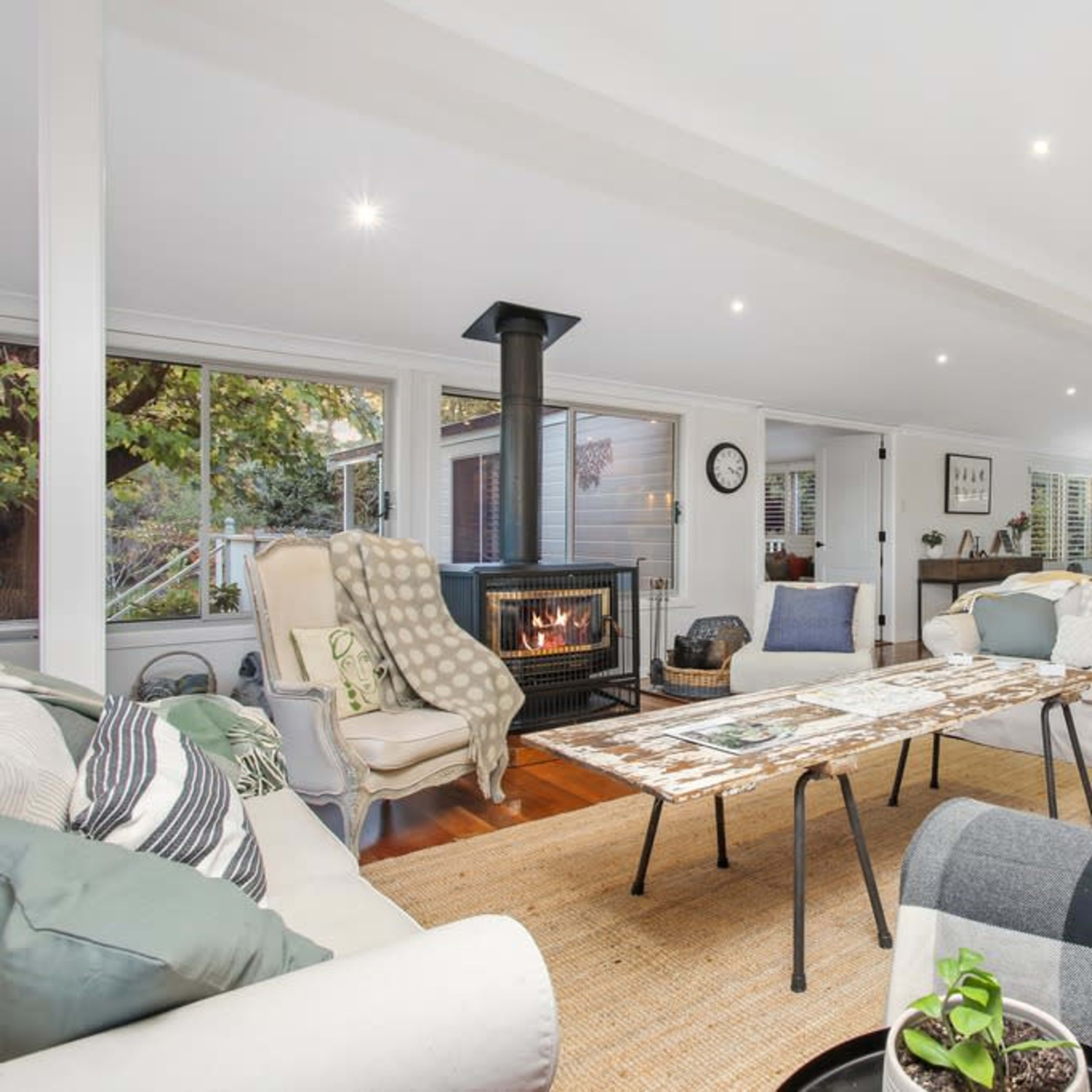 The spacious living room features a central coffee table surrounded by multiple sofas and a fireplace, with large windows offering views of the outdoors.