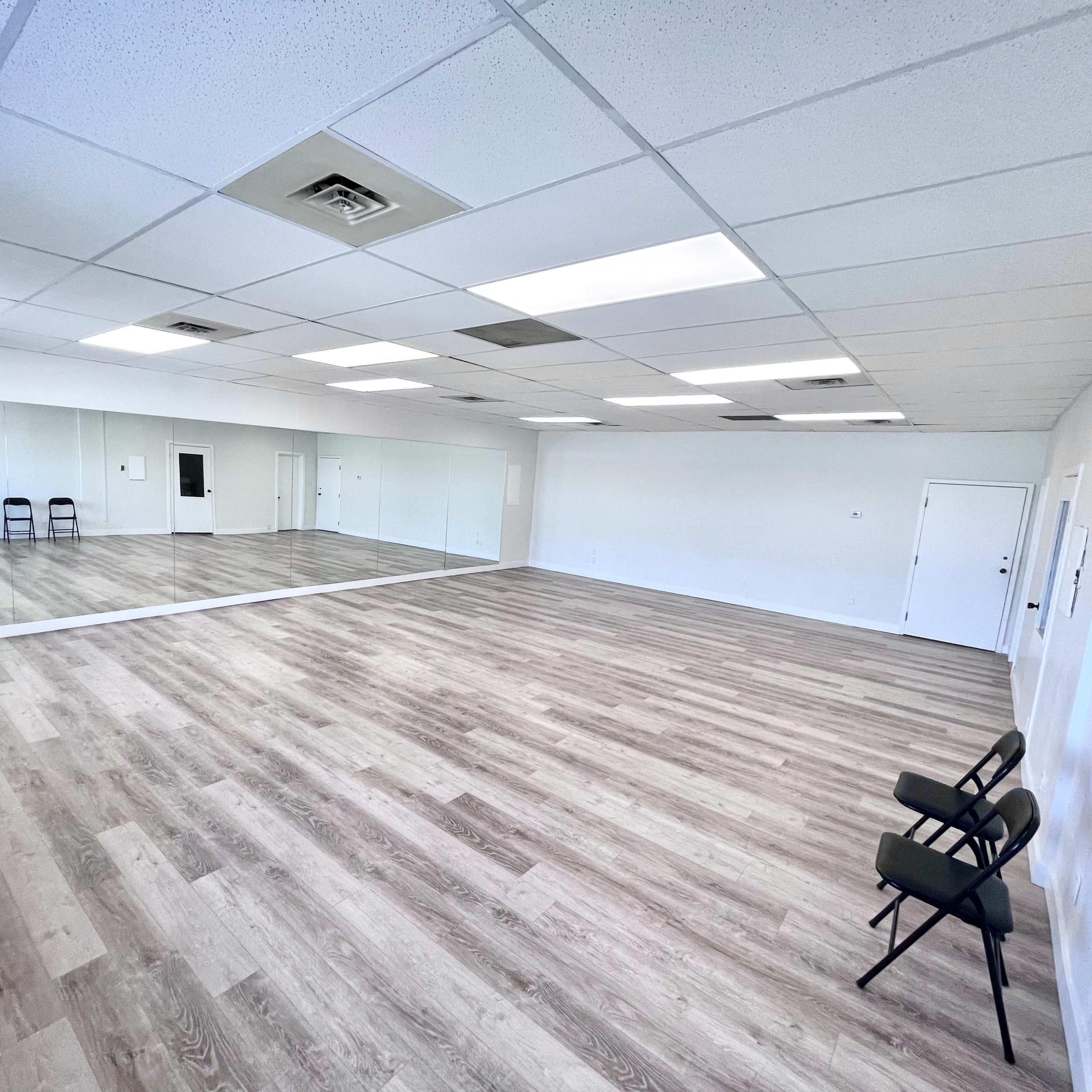 A spacious, empty room with a wooden floor, large mirrors on one wall, and two chairs placed against another wall.