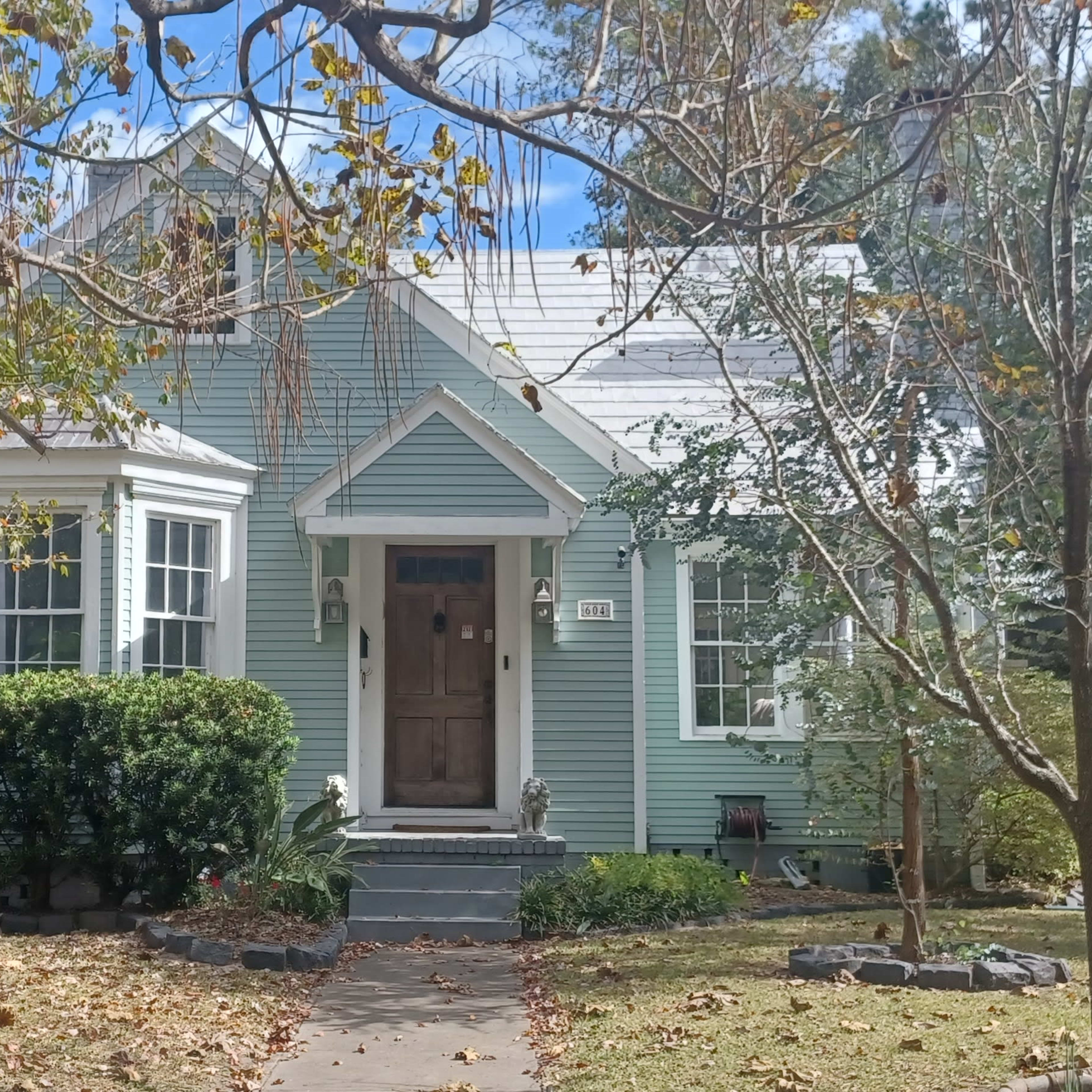 A light green, single-story house with a front porch and decorative windows sits among scattered leaves and trees.