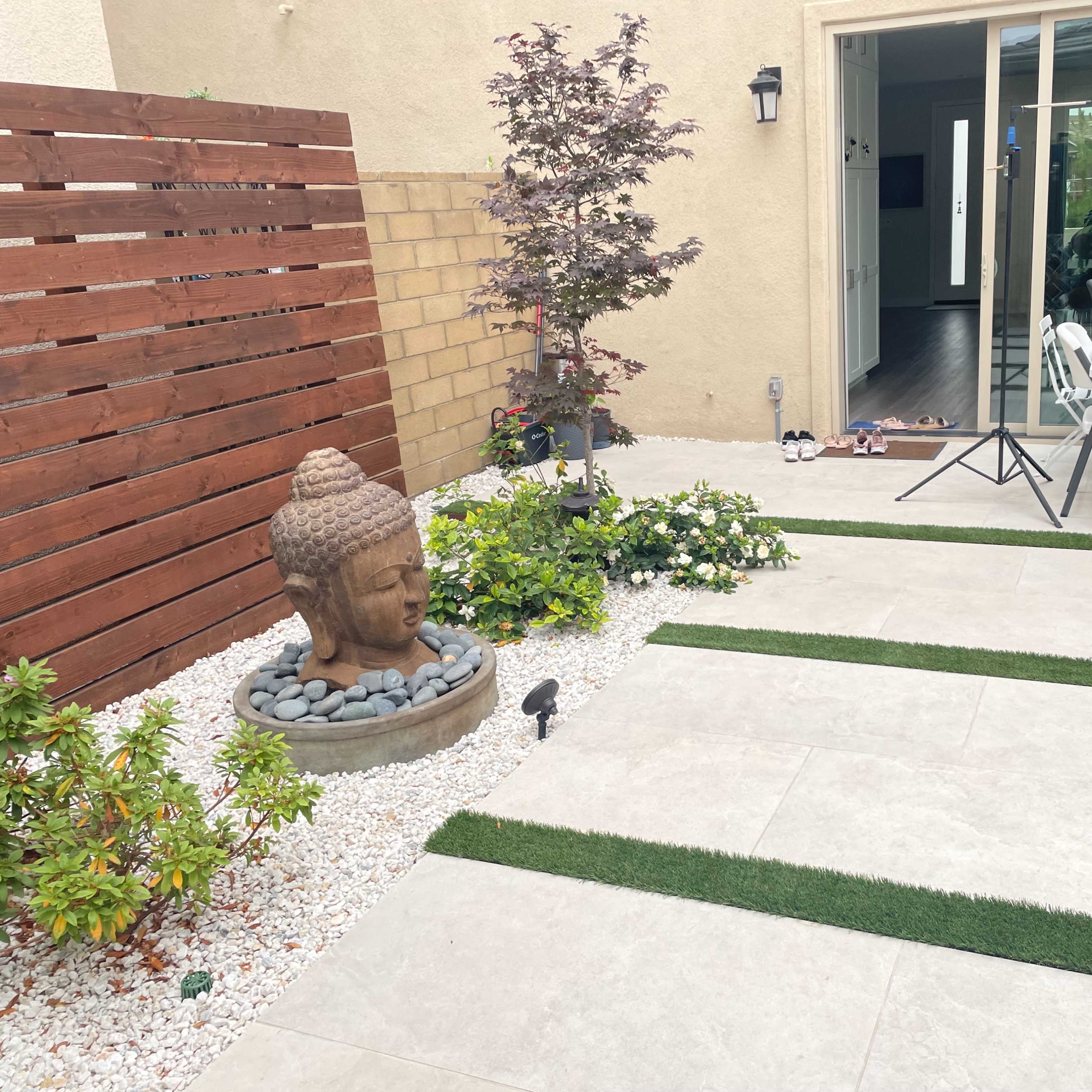A patio area featuring a stone Buddha statue, bordered by plants and decorative gravel, with a small table and chairs visible through a nearby sliding glass door.