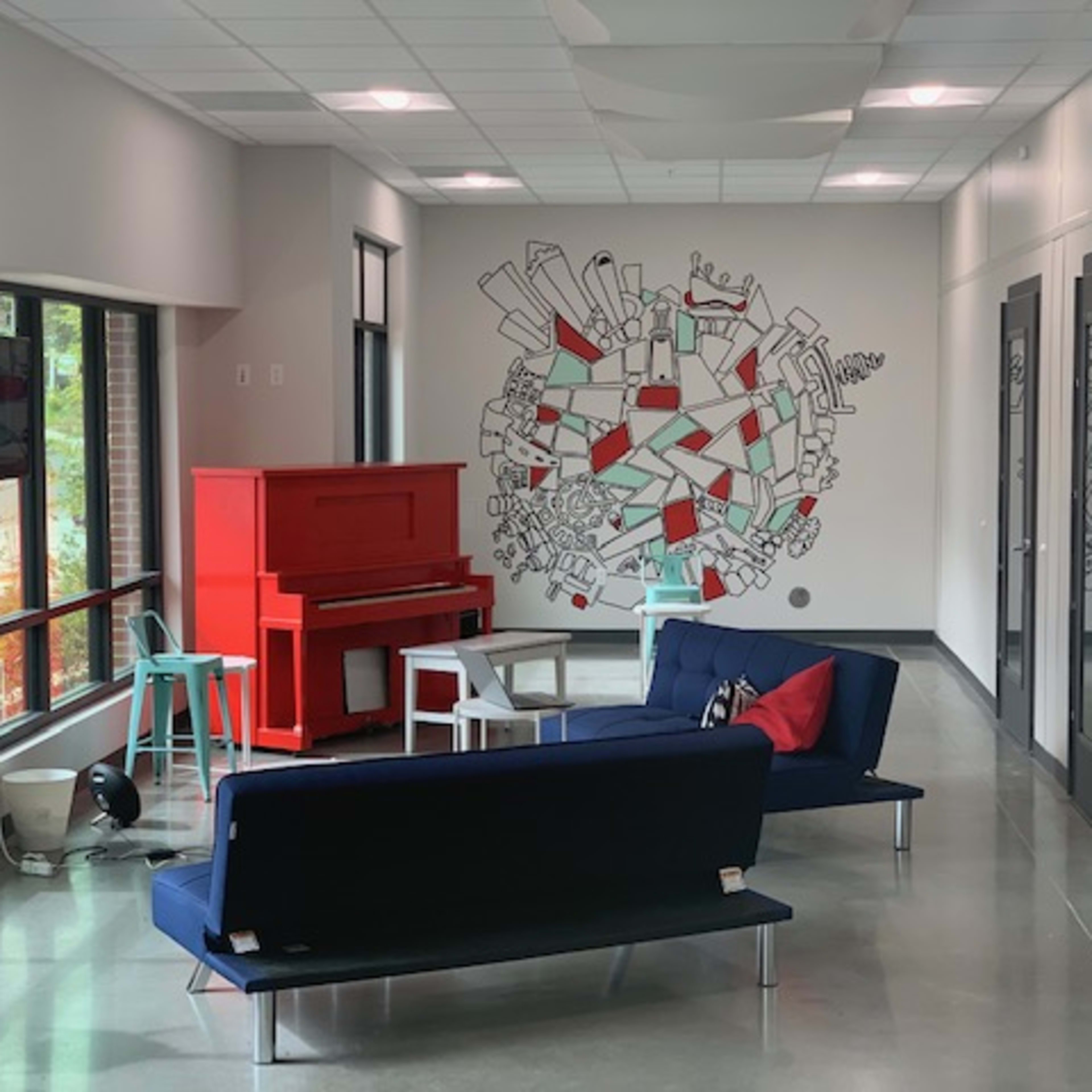 A lounge area features a blue couch, a red piano, and a mural on the wall, with a few chairs and windows allowing natural light.