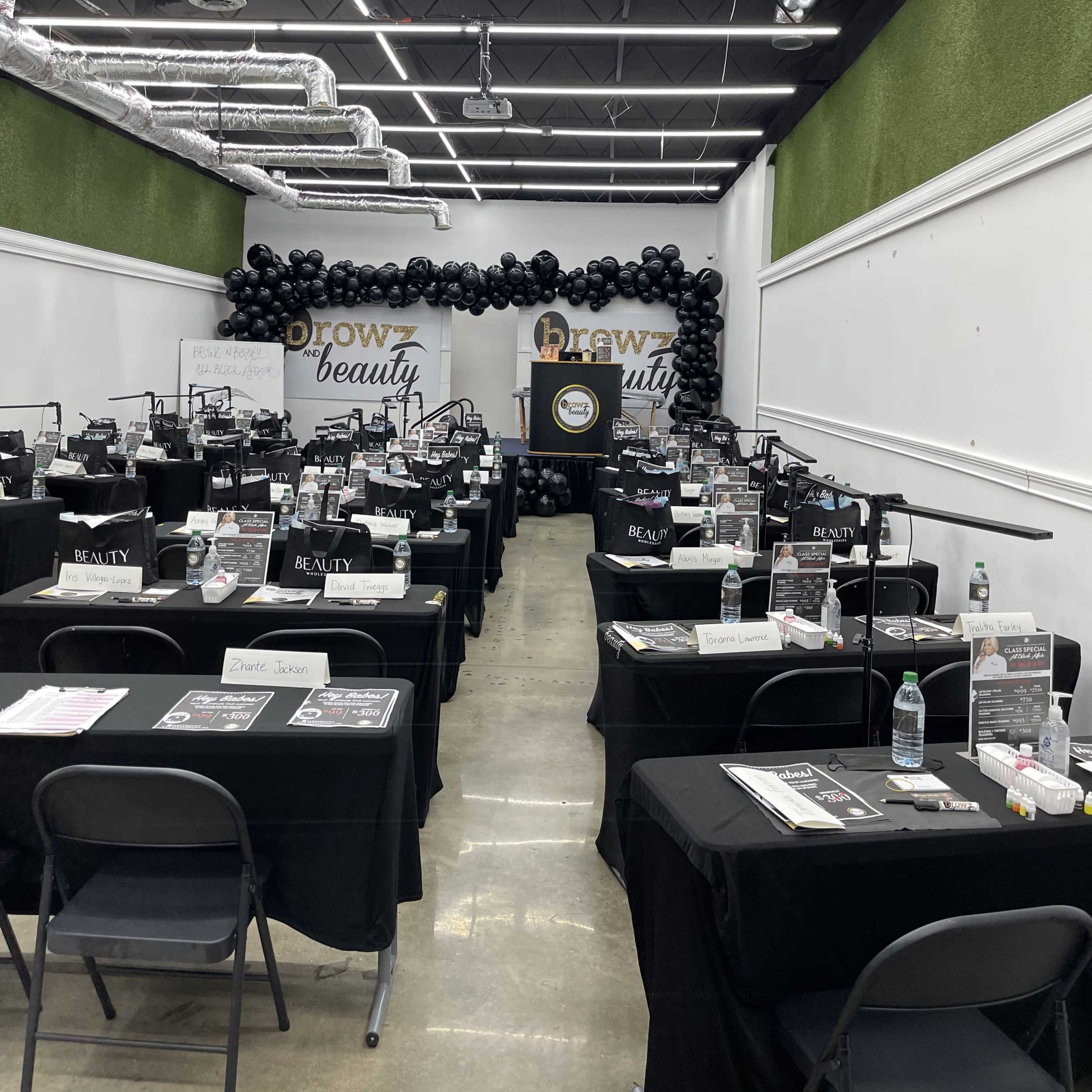 A classroom or event space set up with multiple tables covered in black cloth, each equipped with beauty supplies and tools, arranged in rows under black balloon decorations.