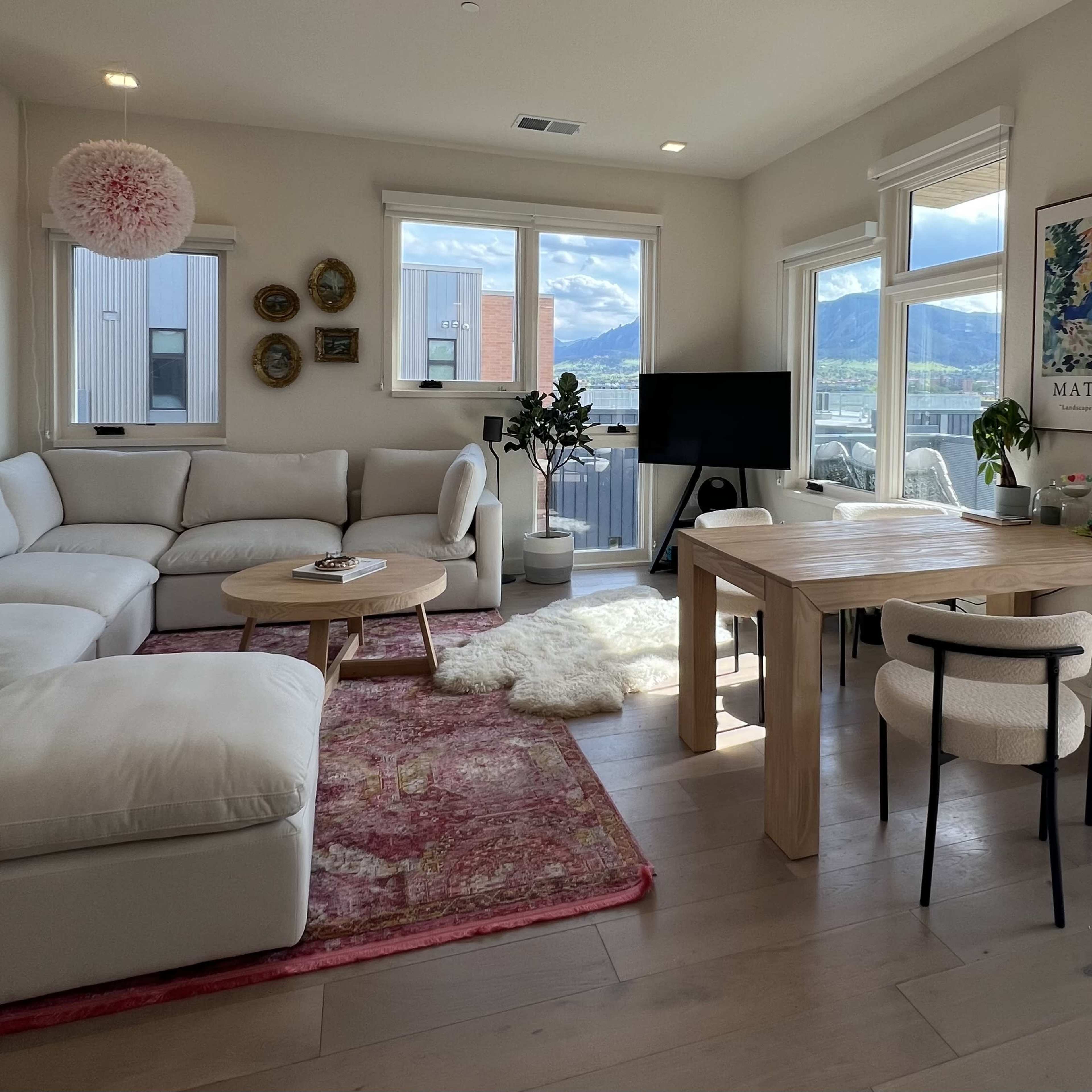 A bright living room features a white sectional couch, a wooden dining table surrounded by chairs, and large windows that offer a view of the outdoors.