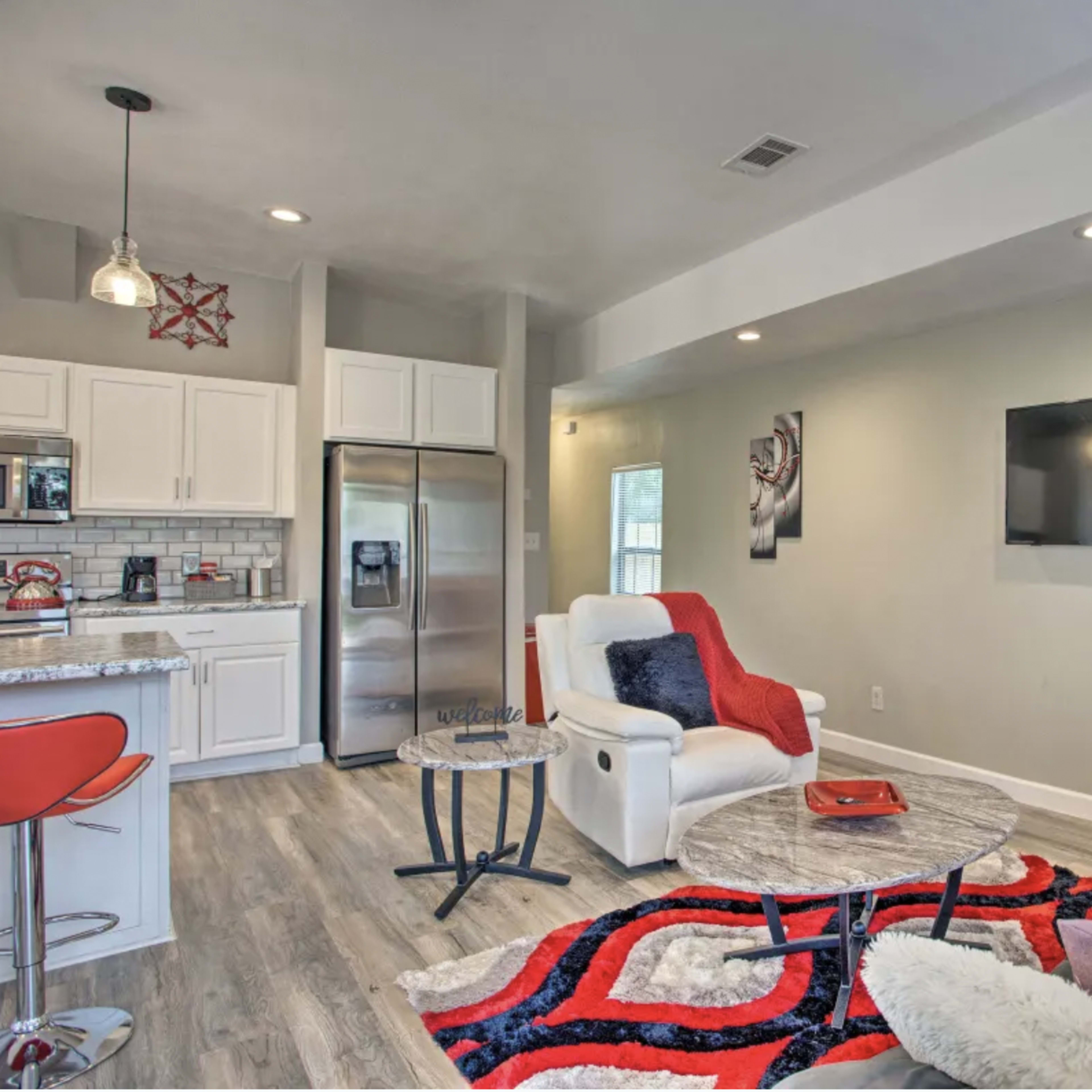 A modern living area featuring a gray sofa with colorful cushions, a coffee table, a wall-mounted television, and a kitchenette with stainless steel appliances.