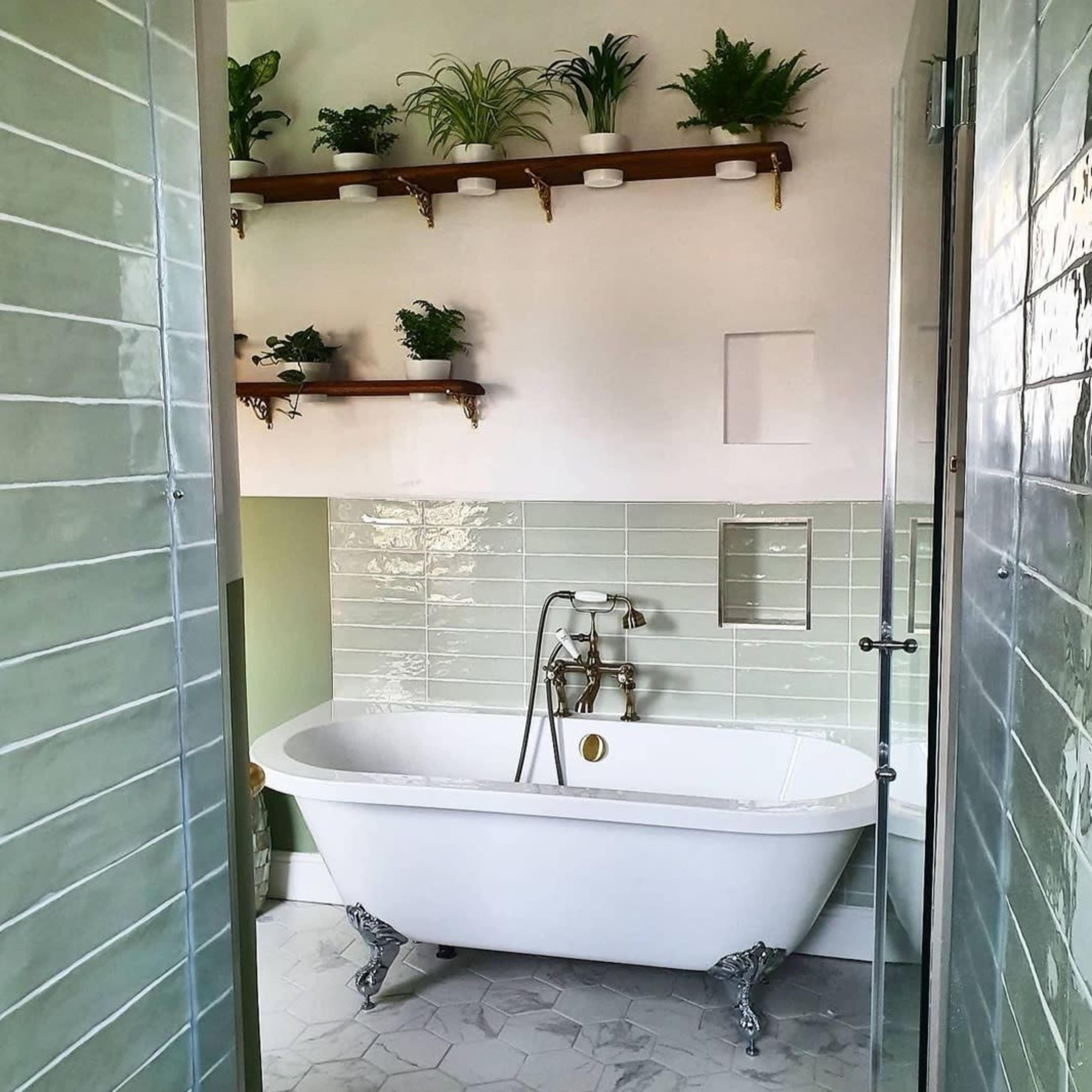 A modern bathroom features a freestanding white bathtub with vintage-style fixtures and green tile walls, complemented by shelves of potted plants.