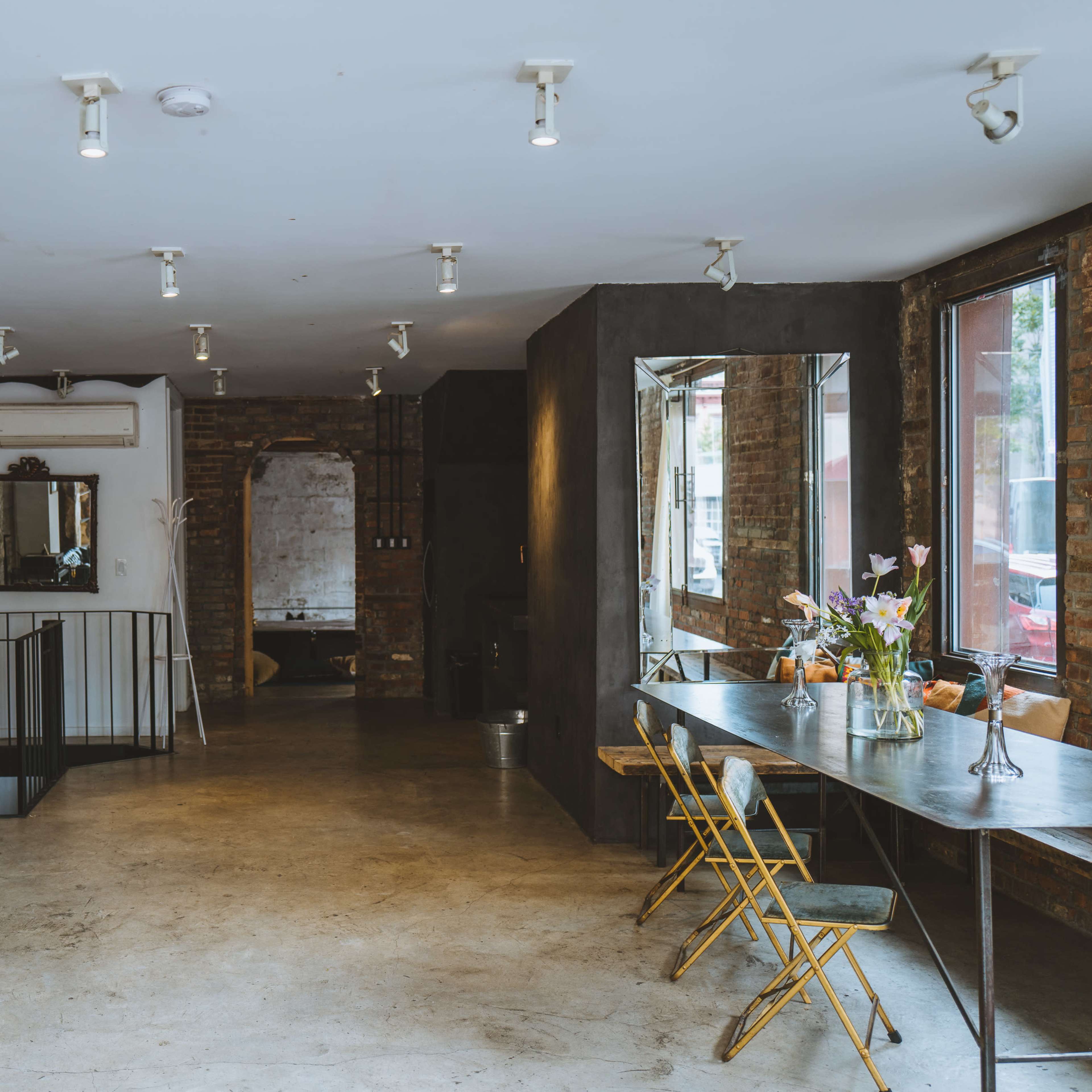 A spacious, modern interior with exposed brick walls, a black leather sofa, a metal dining table, and large windows allowing natural light to enter.