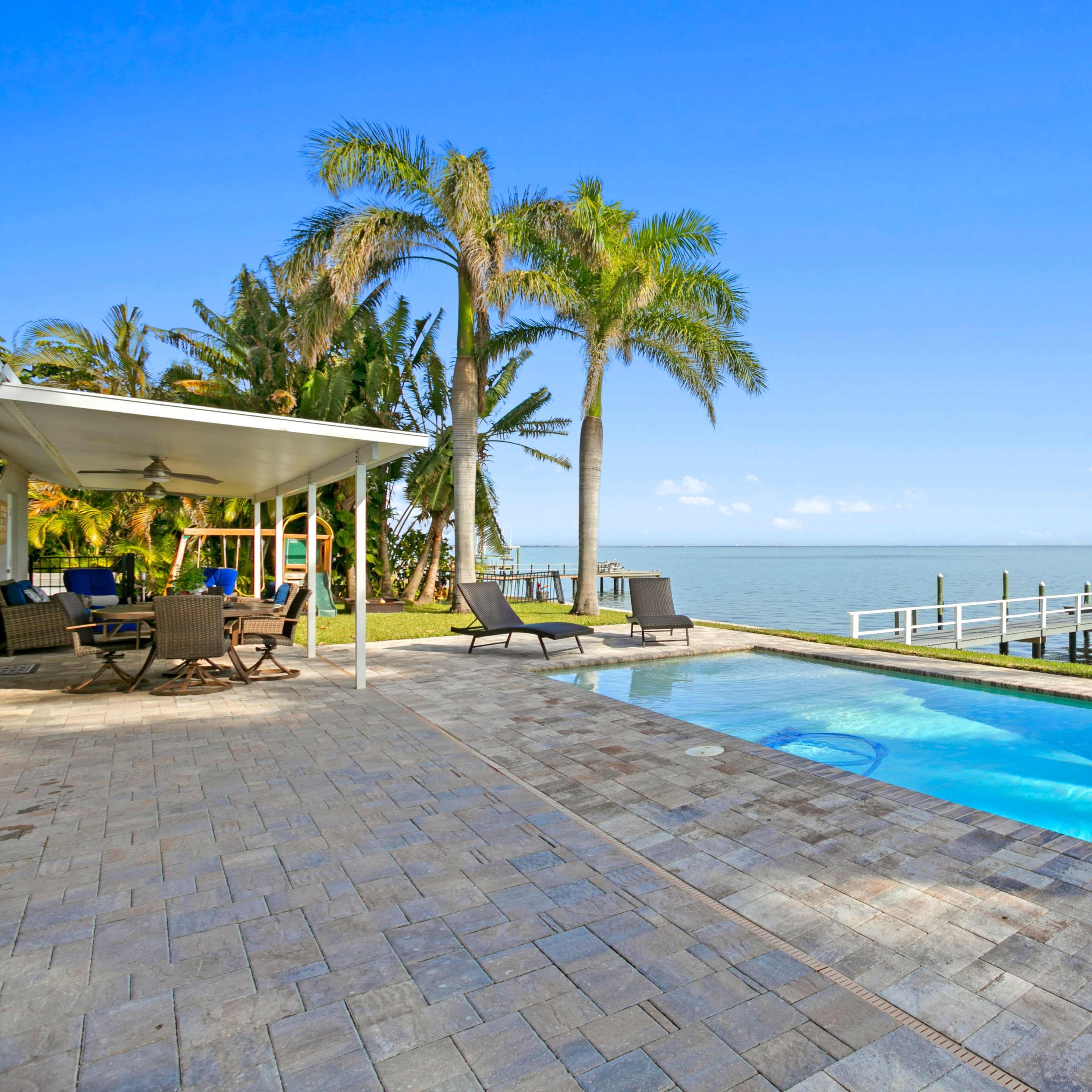 A patio with a swimming pool and a view of the waterfront, featuring palm trees and a dock extending into the water.