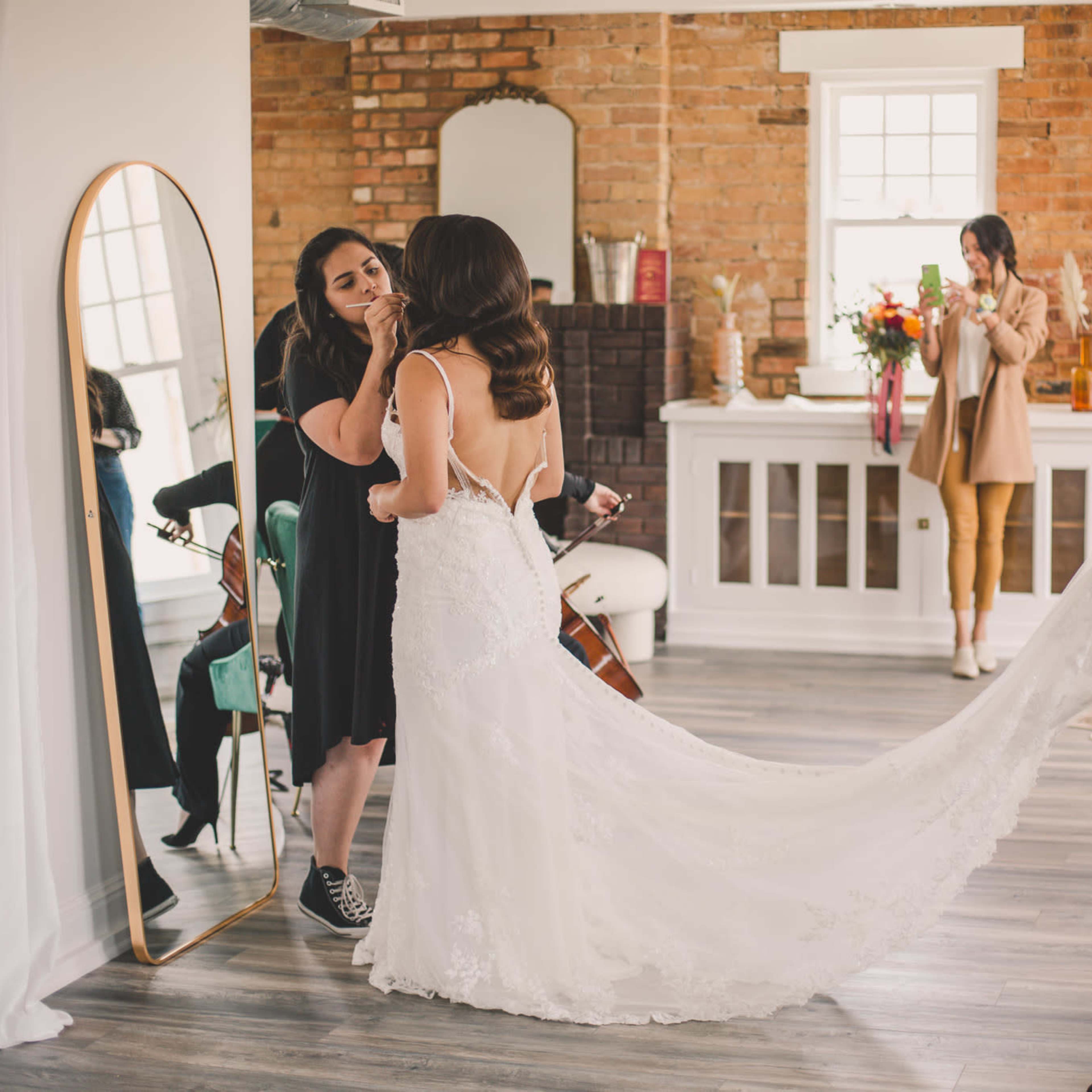 A bride is getting ready for her wedding, assisted by a stylist, while other guests celebrate in a bright, rustic setting.