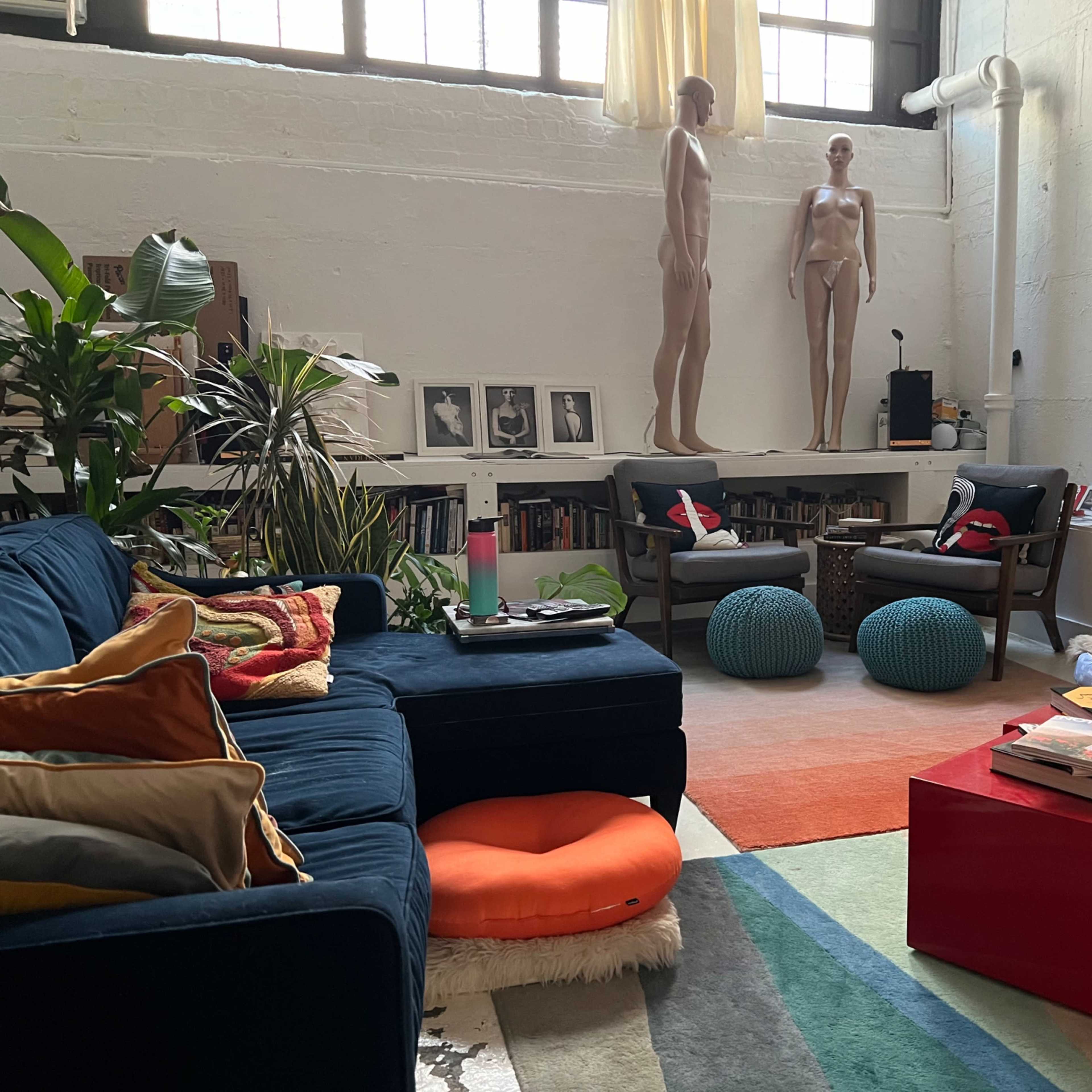 A modern living room with a blue sofa, two mannequin figures, bookshelves, several plants, and colorful rugs arranged on the floor.