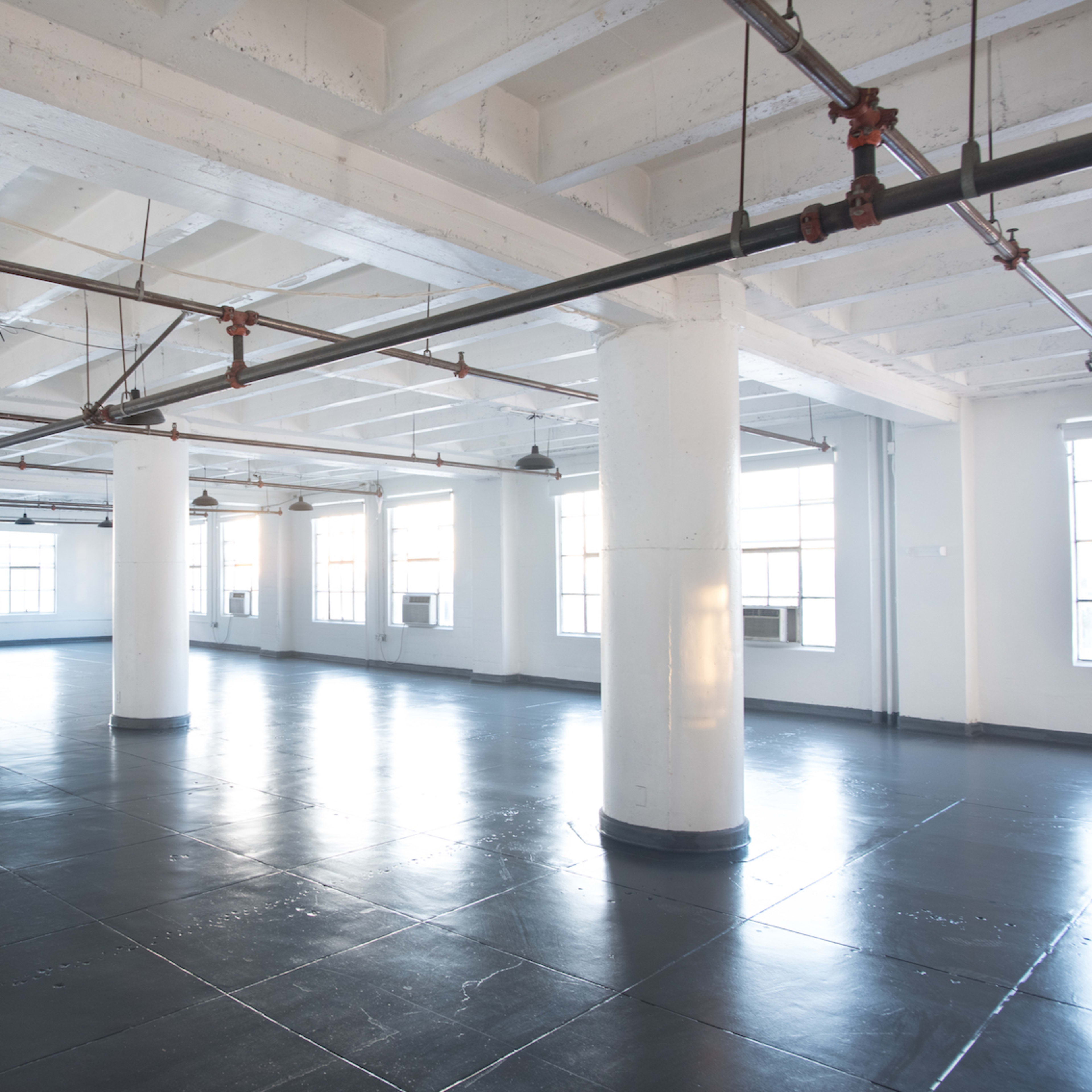A spacious, empty industrial room with polished black floors and beams, featuring white pillars and large windows.