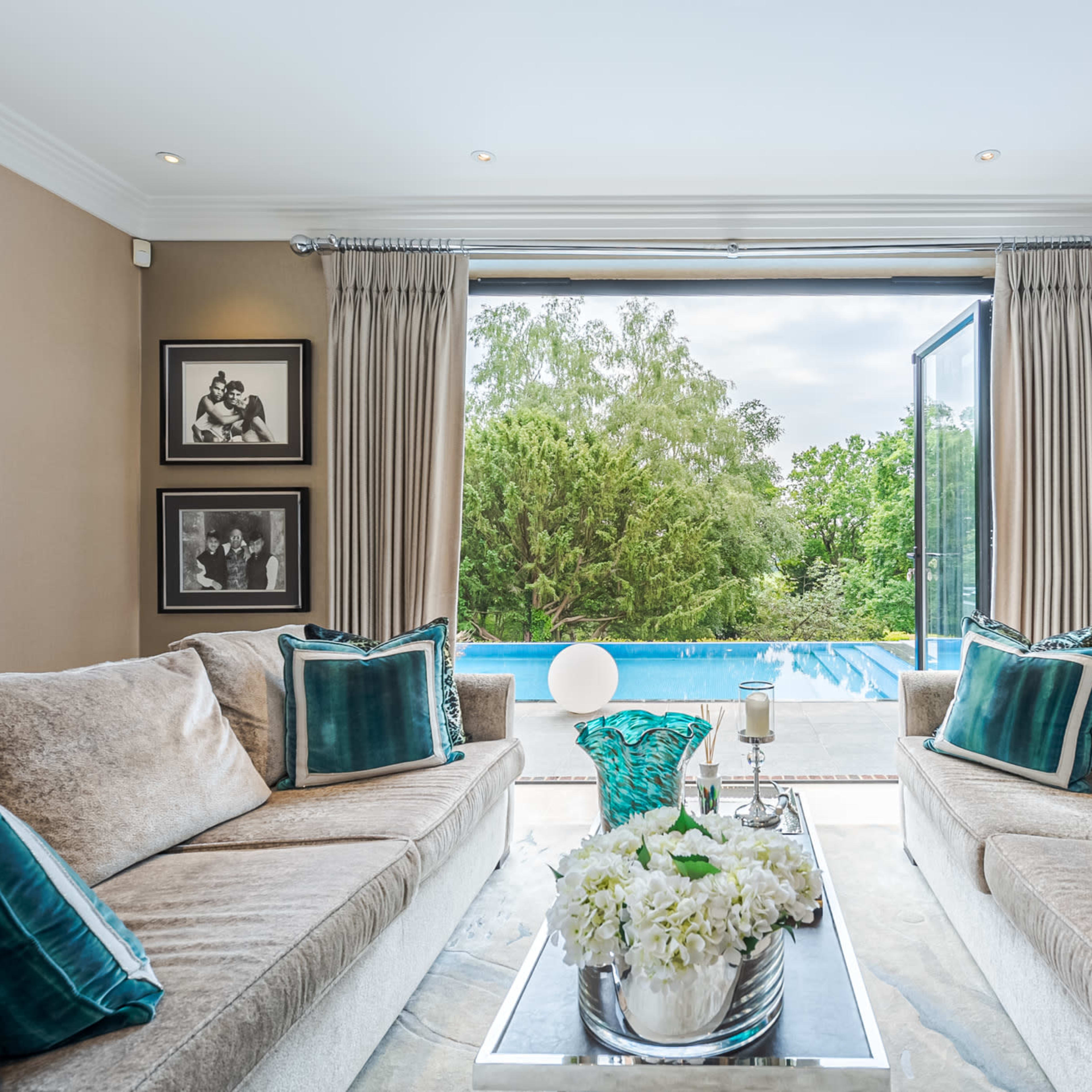 The living room features two sofas with teal accent pillows, a glass coffee table, and large windows opening to a poolside view.