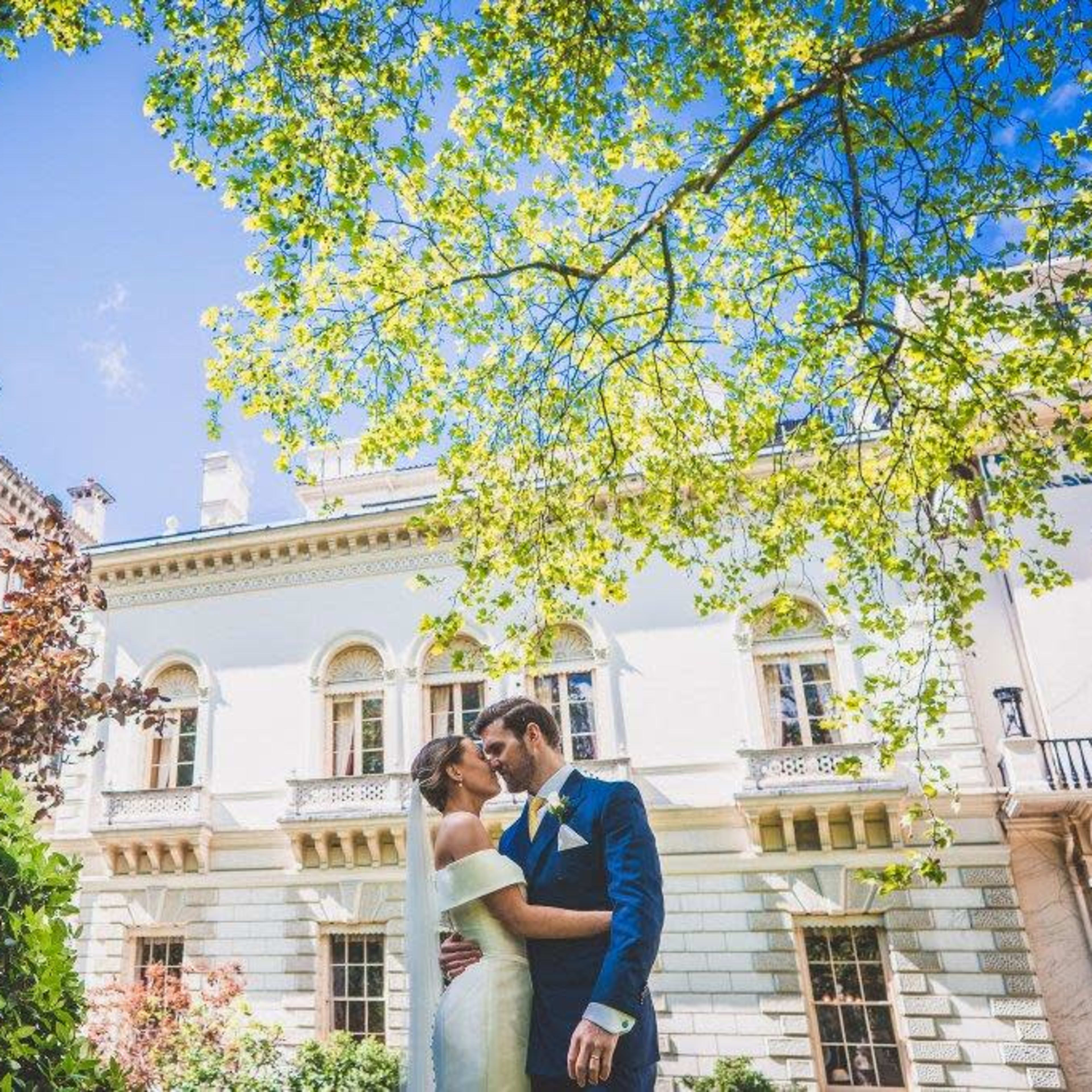 A bride and groom kiss in a lush garden in front of an elegant mansion.