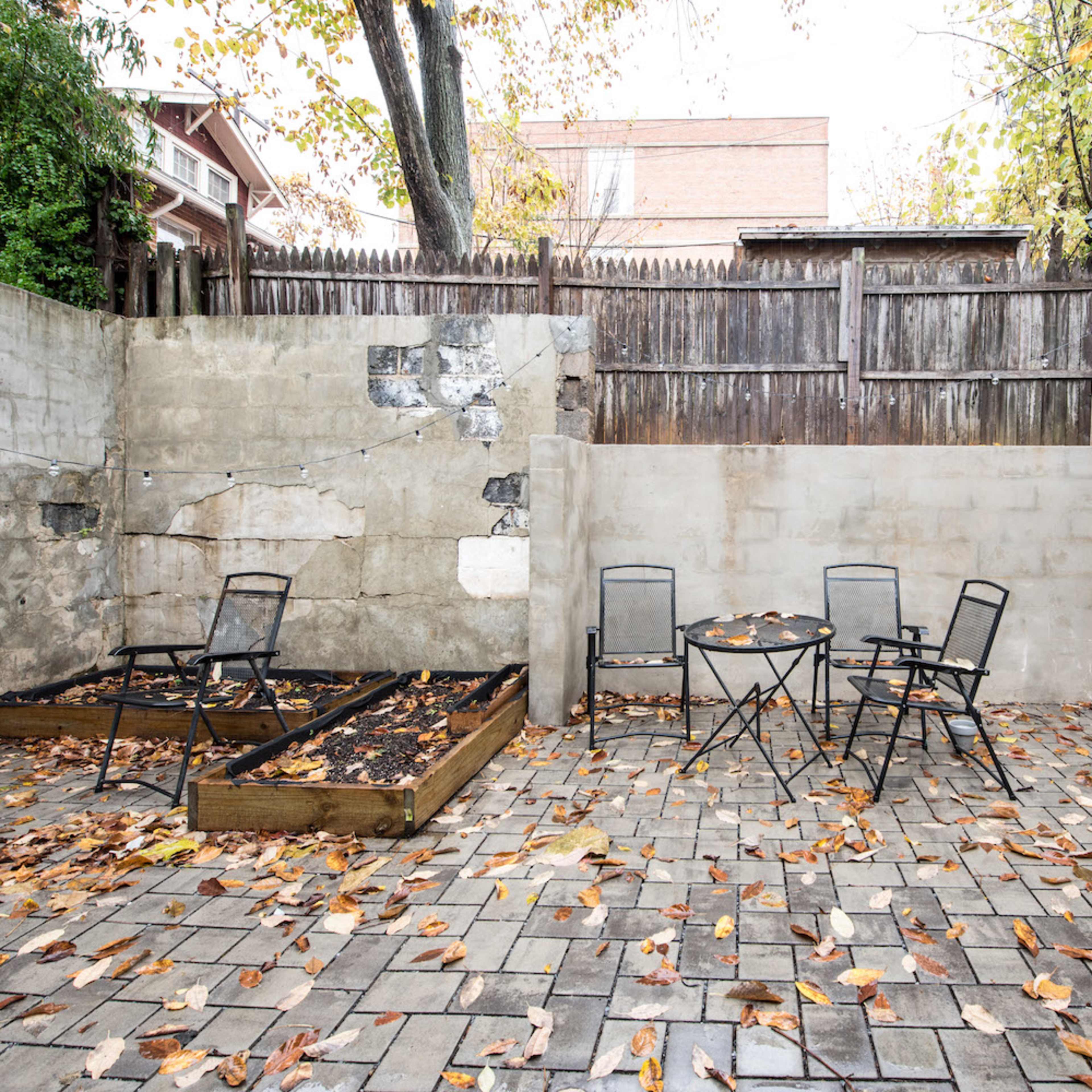 A small, landscaped patio area with two metal chairs and a round table surrounded by fallen leaves and a raised garden bed.