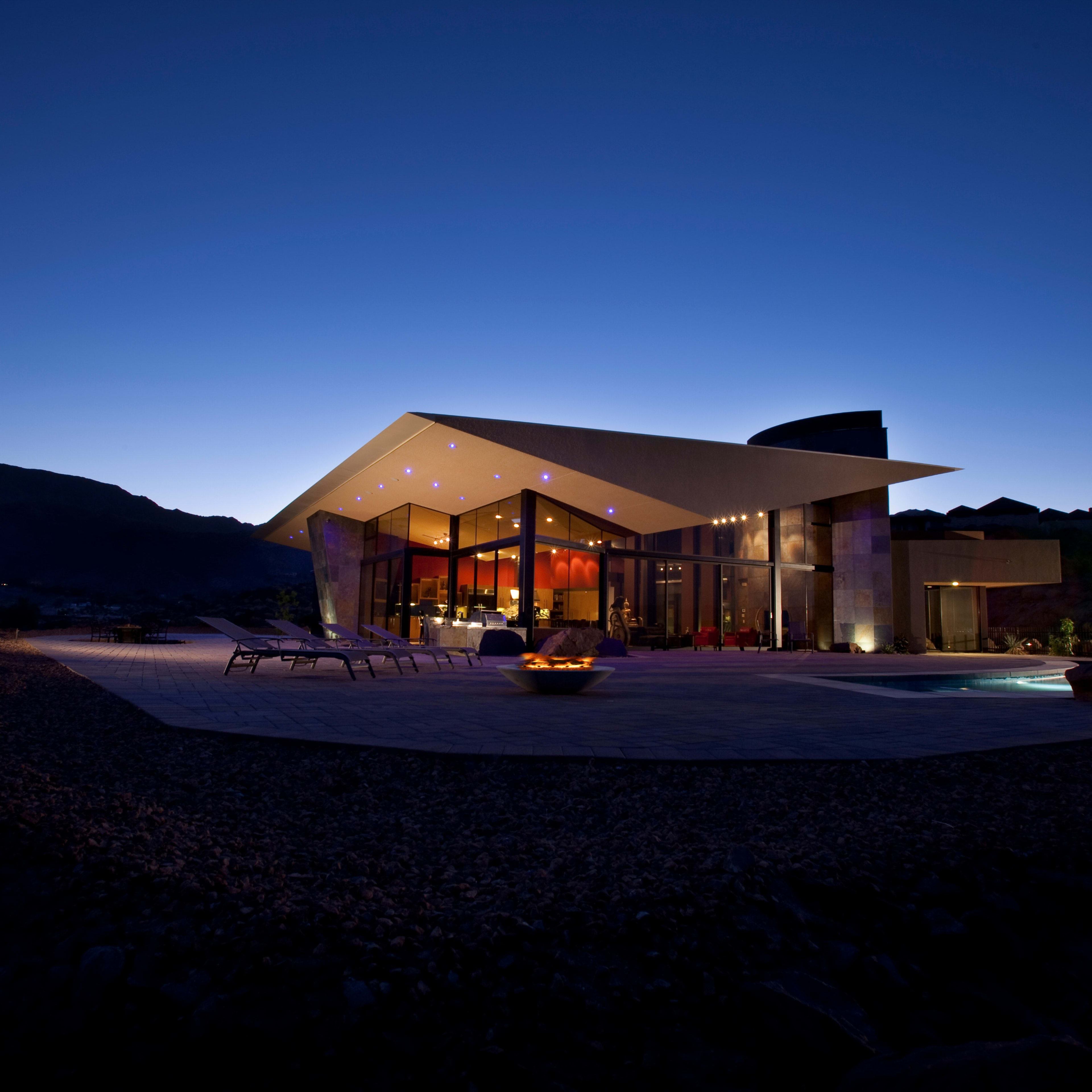 A modern architectural villa with large glass windows and a flat roof, illuminated at twilight against a mountainous backdrop.