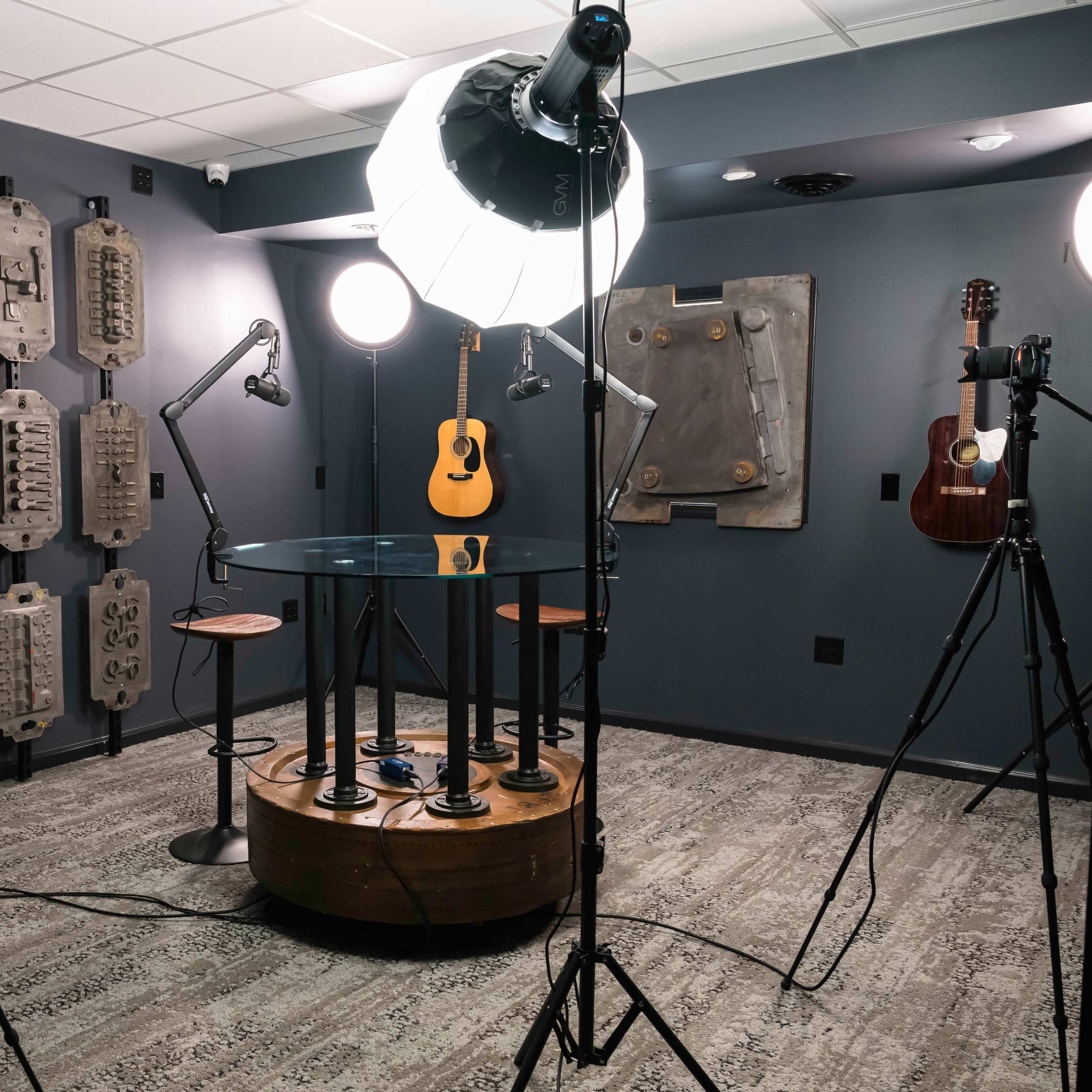 A photography studio with a circular glass table, two guitars mounted on the wall, and multiple cameras set up on tripods under soft lighting.