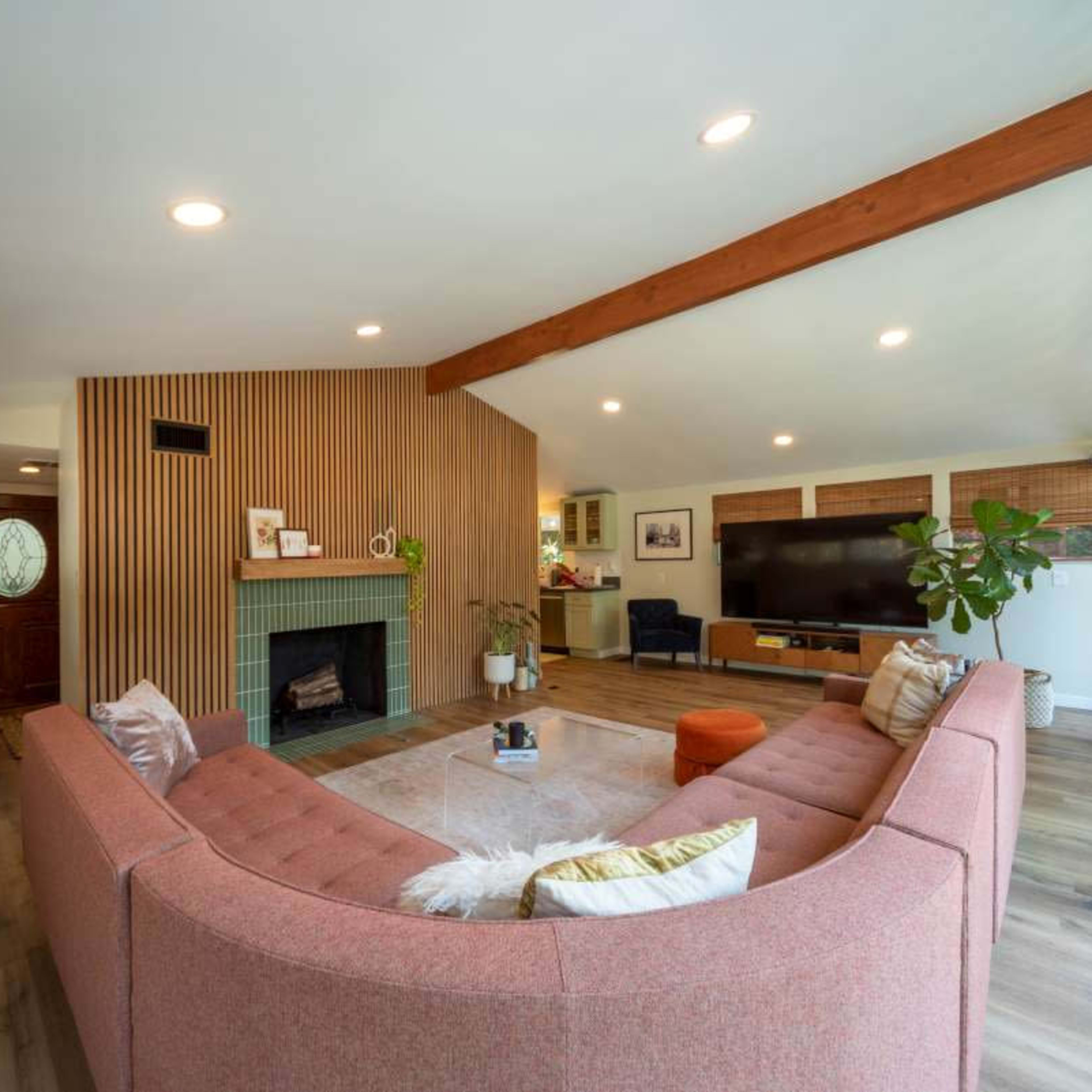 A modern living room featuring a pink sectional sofa, a fireplace with wooden paneling, and large windows with natural light.