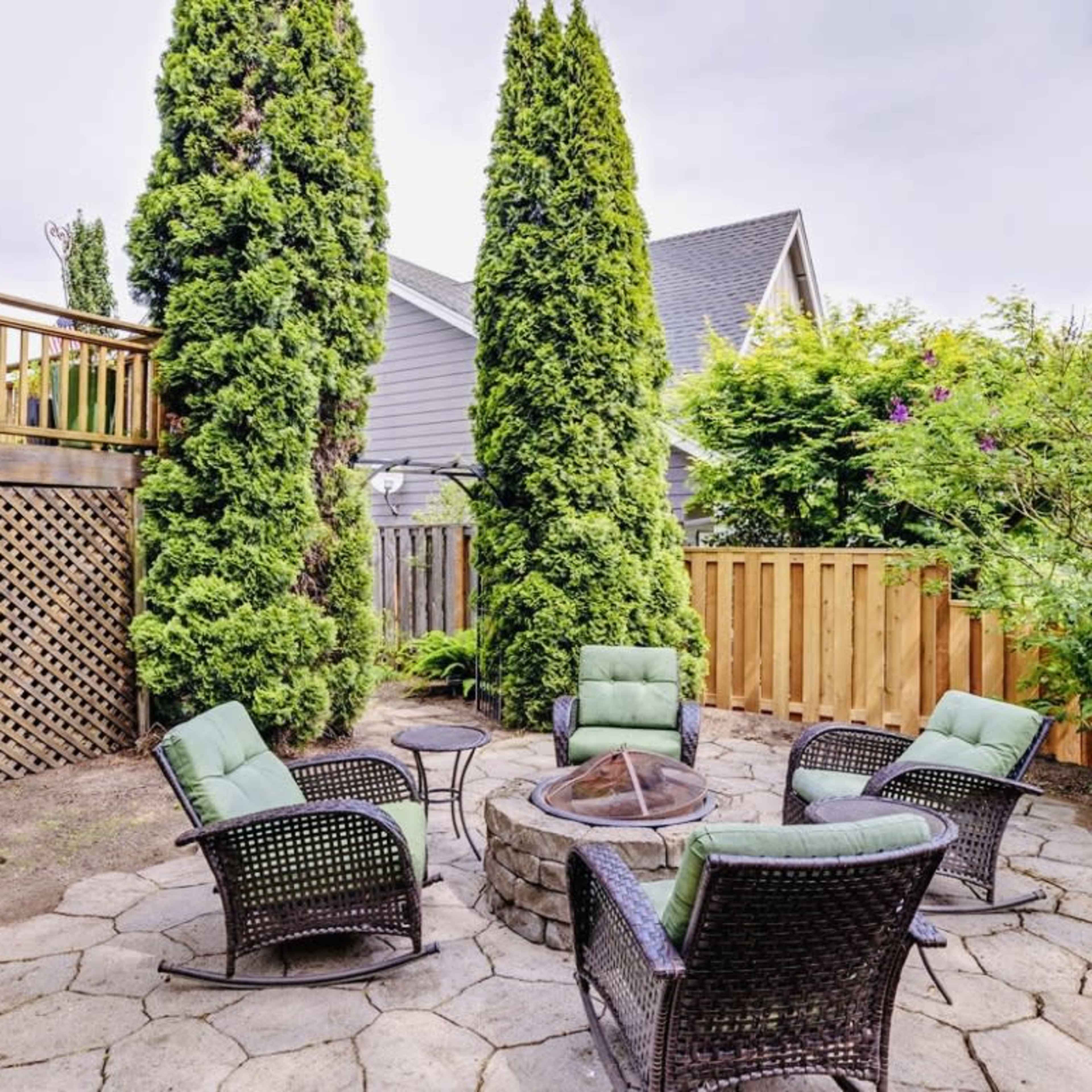 A circular seating area with four chairs surrounds a fire pit, bordered by tall trees and a wooden fence.