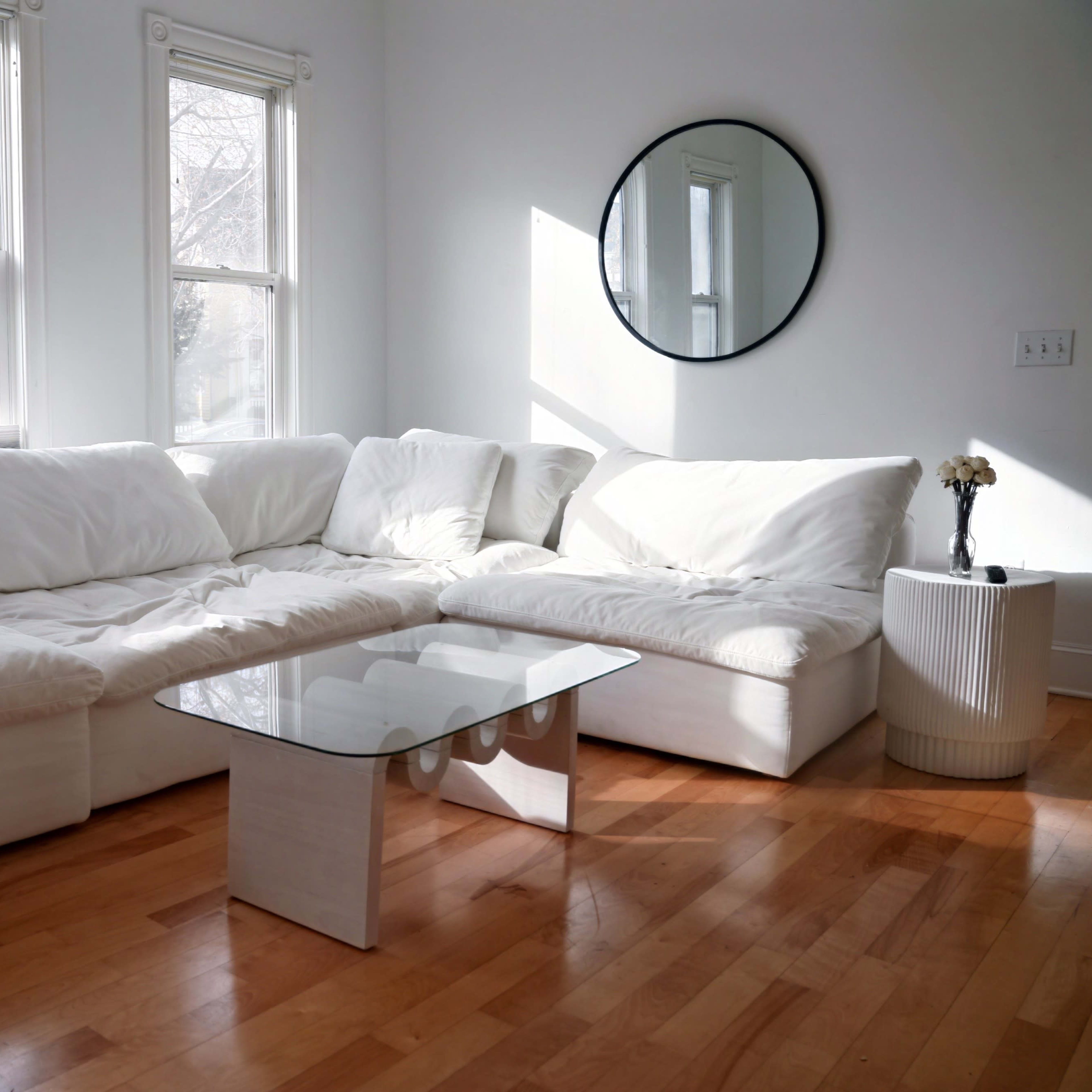 A bright room with a white sectional sofa, a glass coffee table, and large windows allowing natural light to enter.