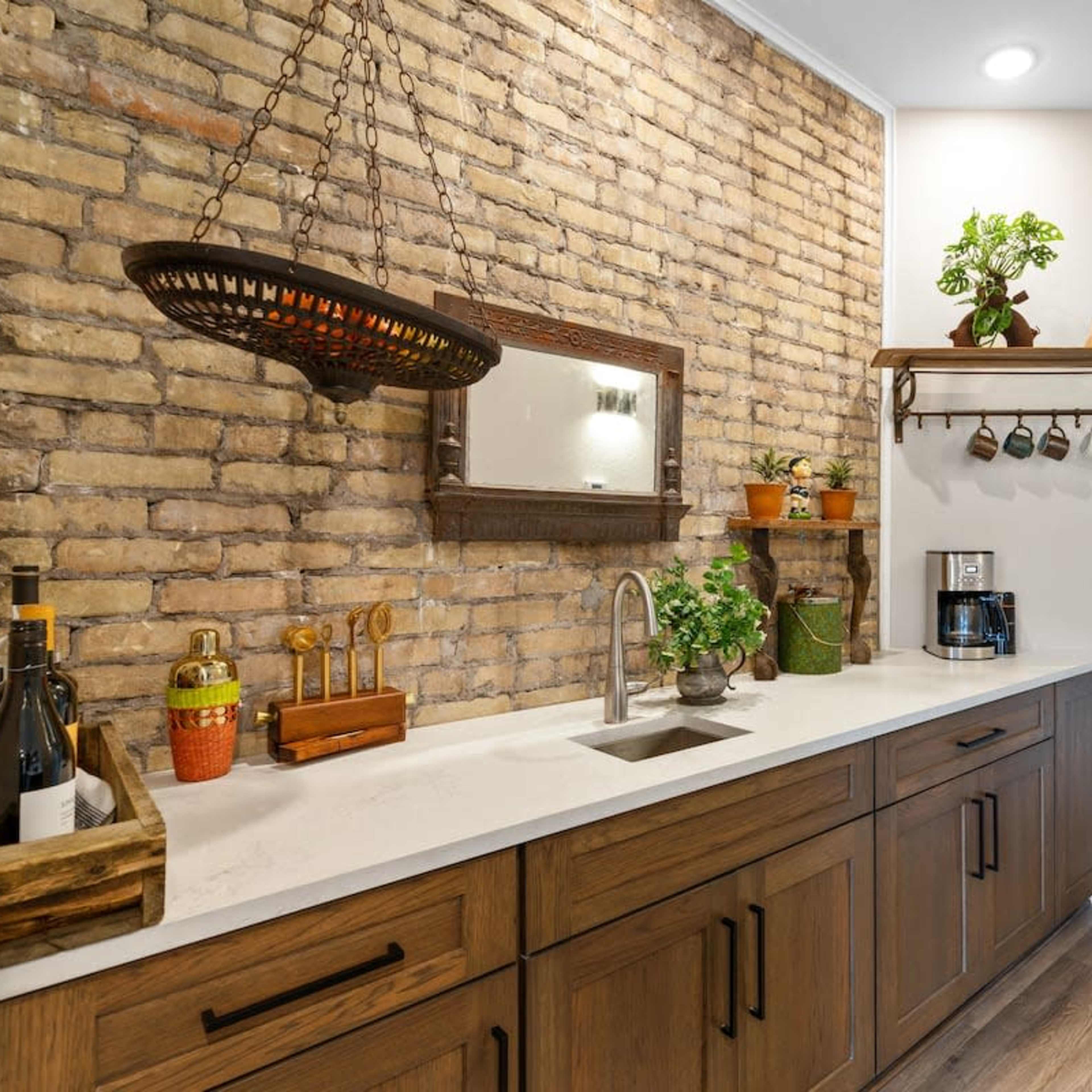 A kitchen space with a brick accent wall, wooden cabinetry, a sink, and various potted plants and kitchen items arranged on the counter.