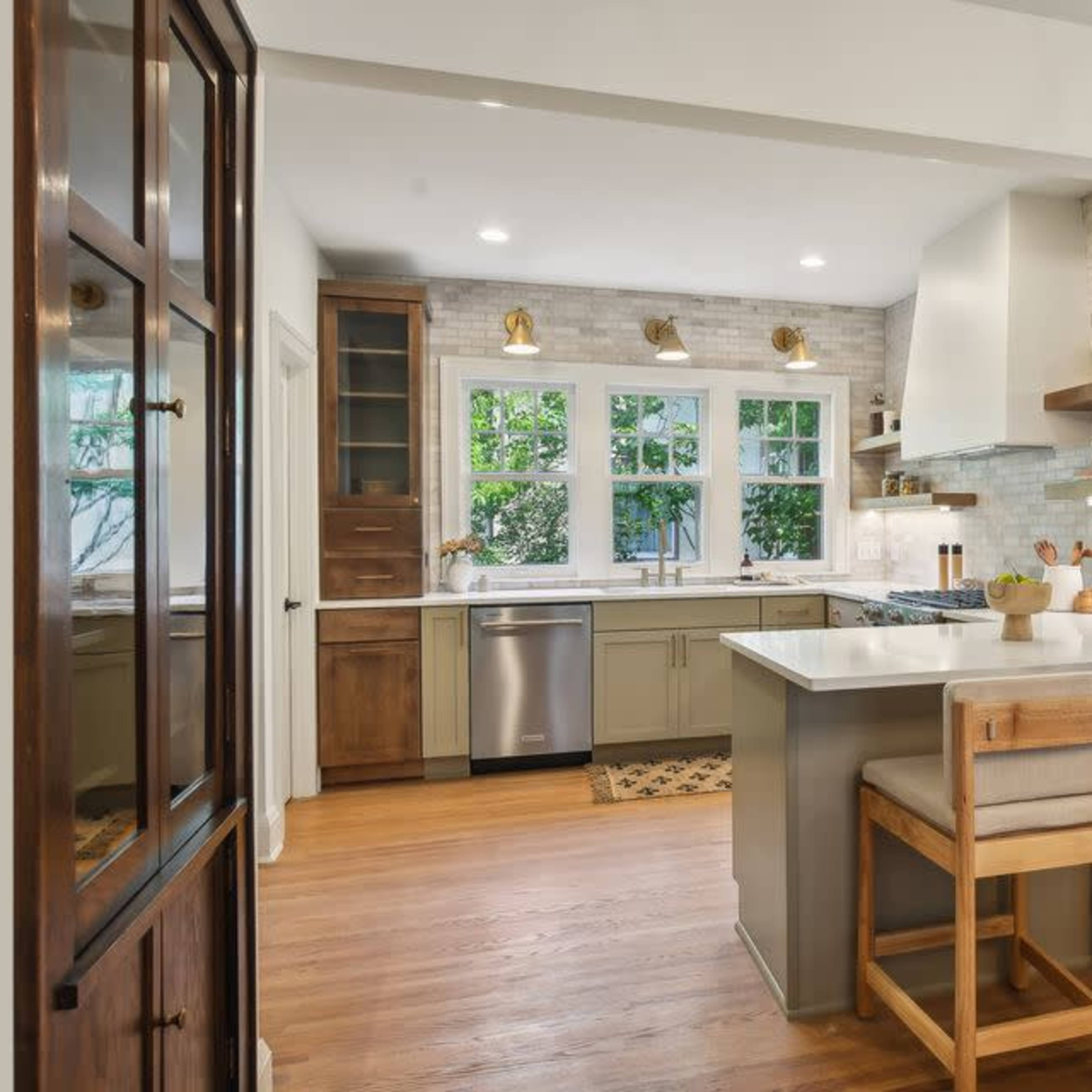 A modern kitchen features wooden cabinets, stainless steel appliances, and a central island with two stools, illuminated by natural light from the windows.