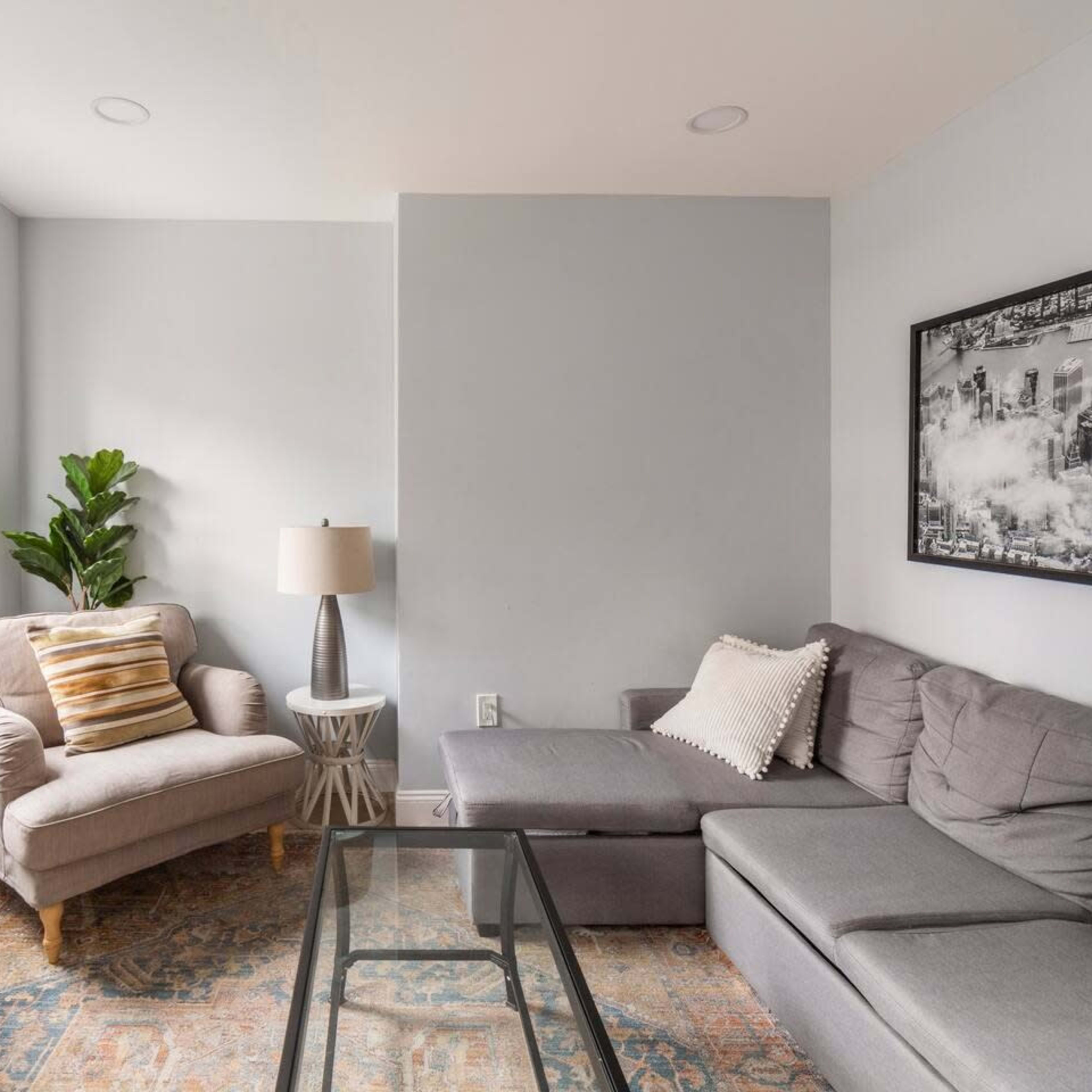 The room features a gray sectional sofa, a cozy armchair with a striped pillow, a glass coffee table, and a television mounted on the wall.