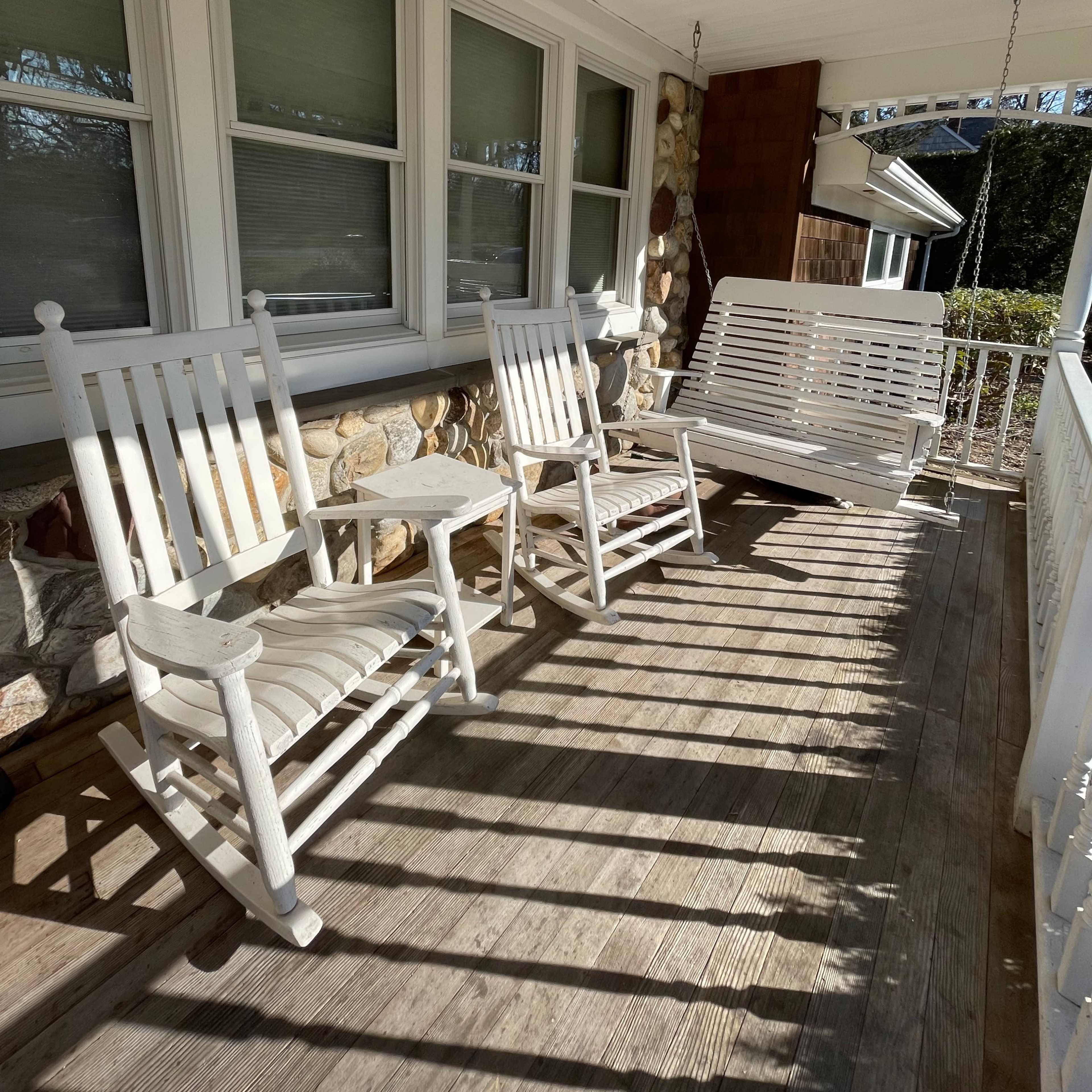 A porch with two white rocking chairs and a white swing, positioned on a wooden floor and surrounded by stone walls.