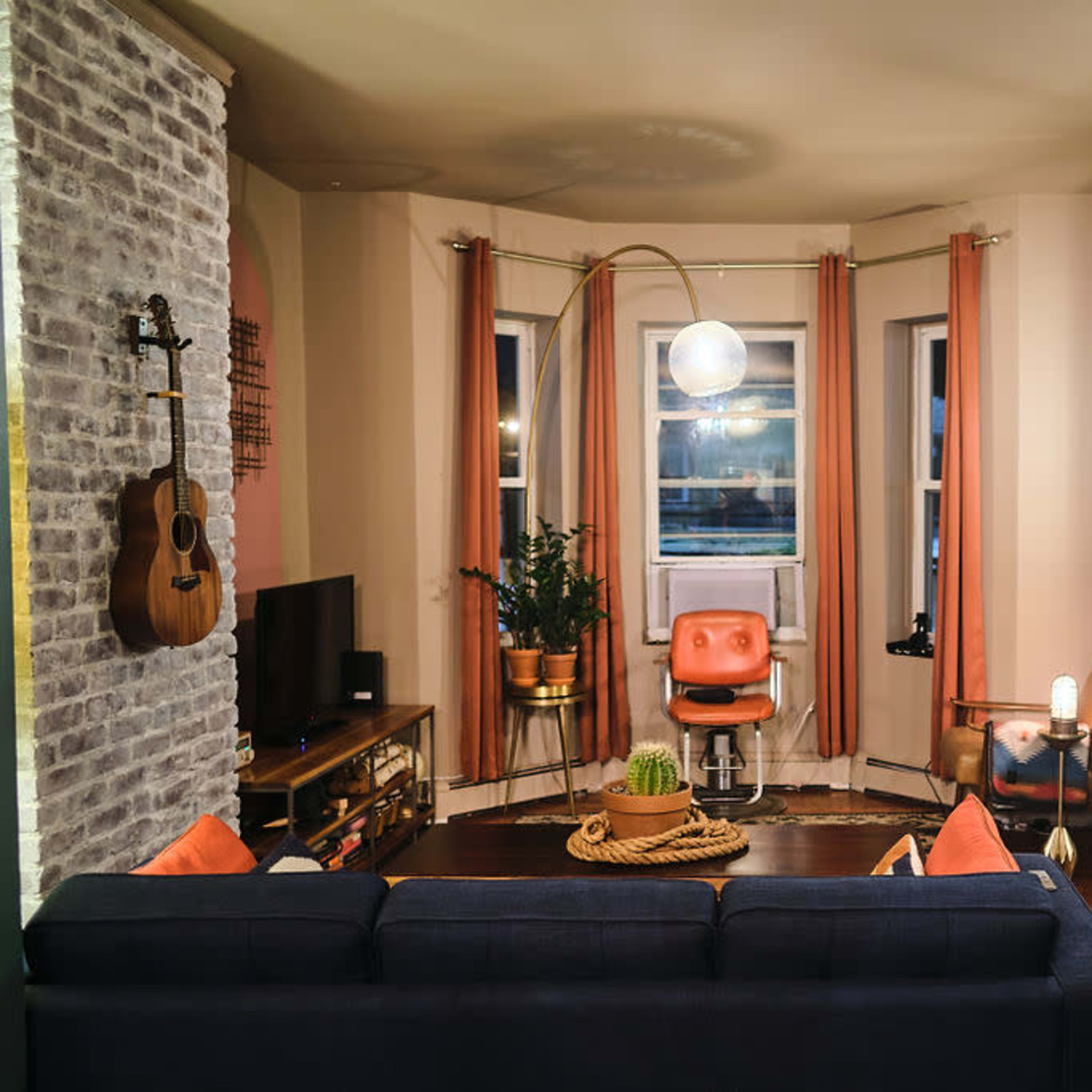 A cozy living room featuring a sofa, a guitar hanging on the wall, a small table with a lamp, and large windows dressed with curtains.