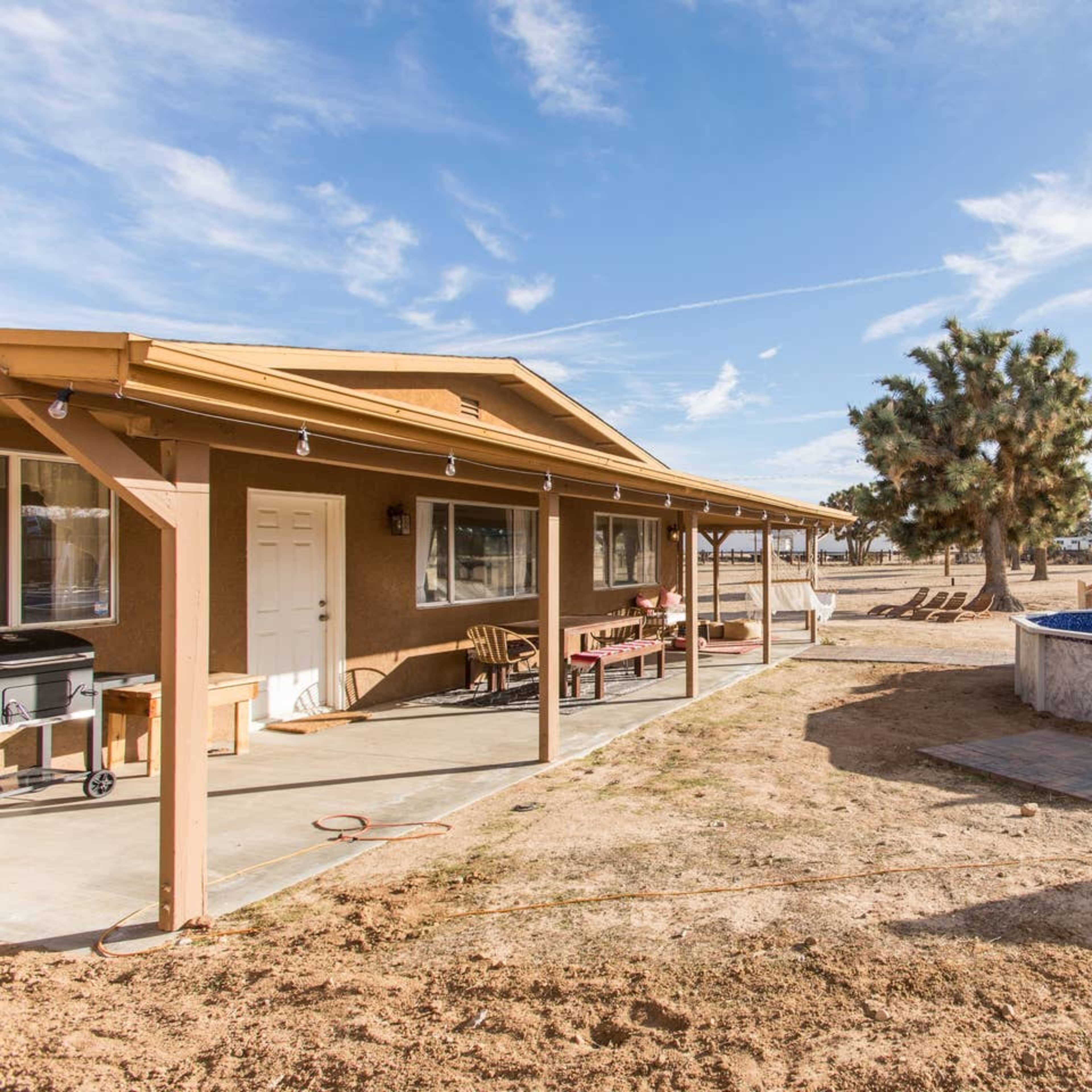 A house with a covered porch and a round above-ground pool is situated in a desert landscape with sparse vegetation and clear blue skies.