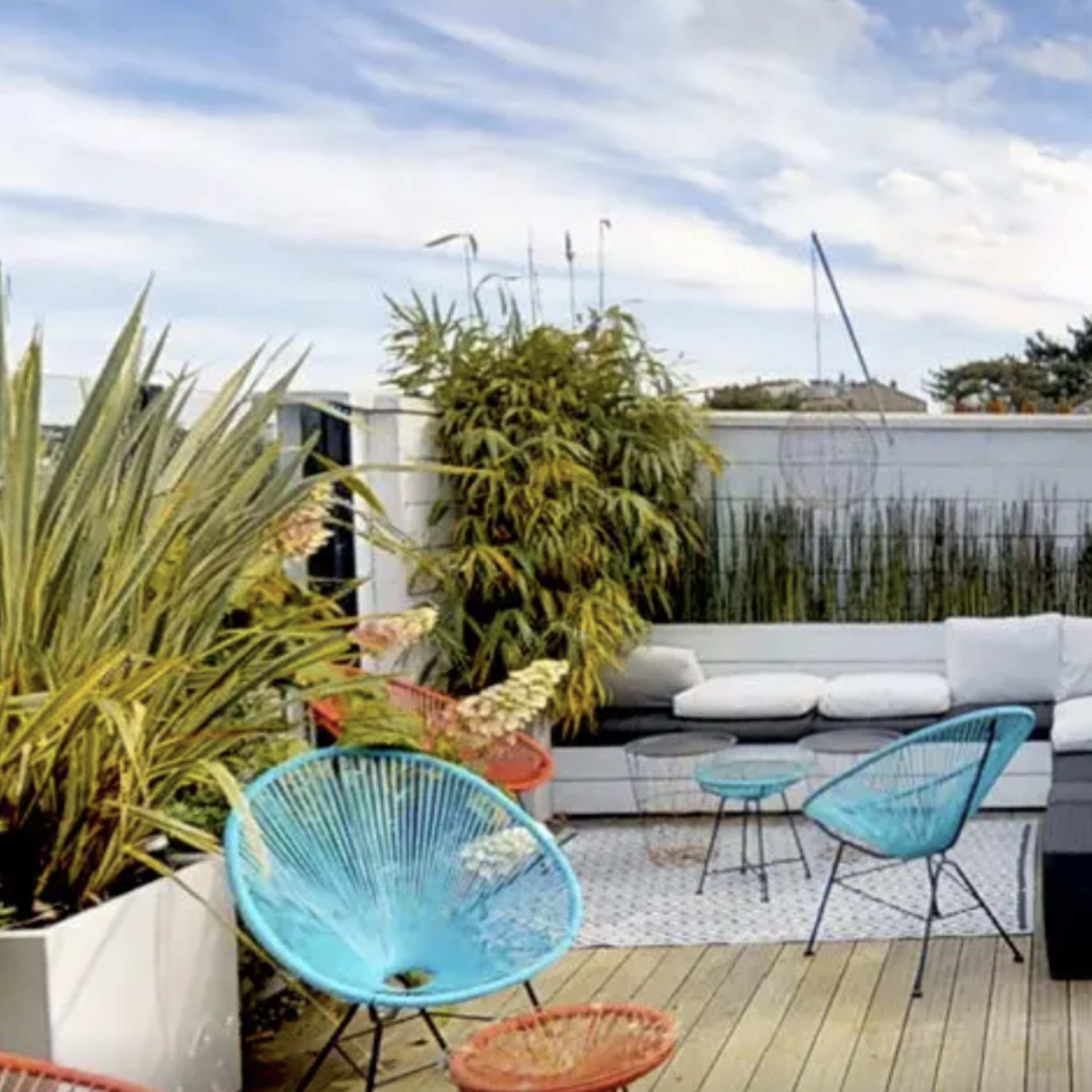 A modern rooftop terrace with a lounge area featuring a white sofa, colorful chairs, and various plants, surrounded by a wooden deck.