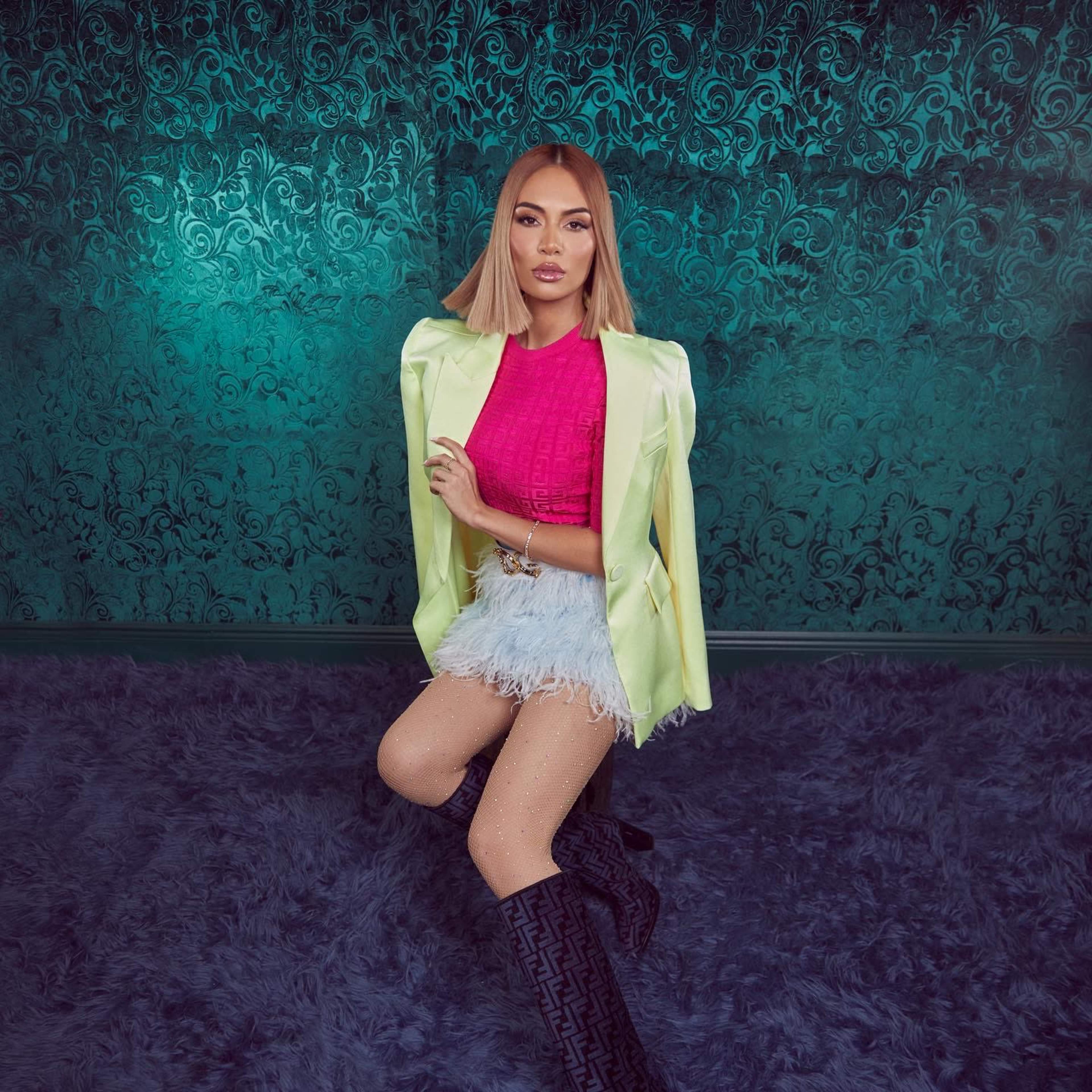 A person stands confidently in a stylish pink top, a pastel green blazer, and a feathered skirt against a decorative teal background.