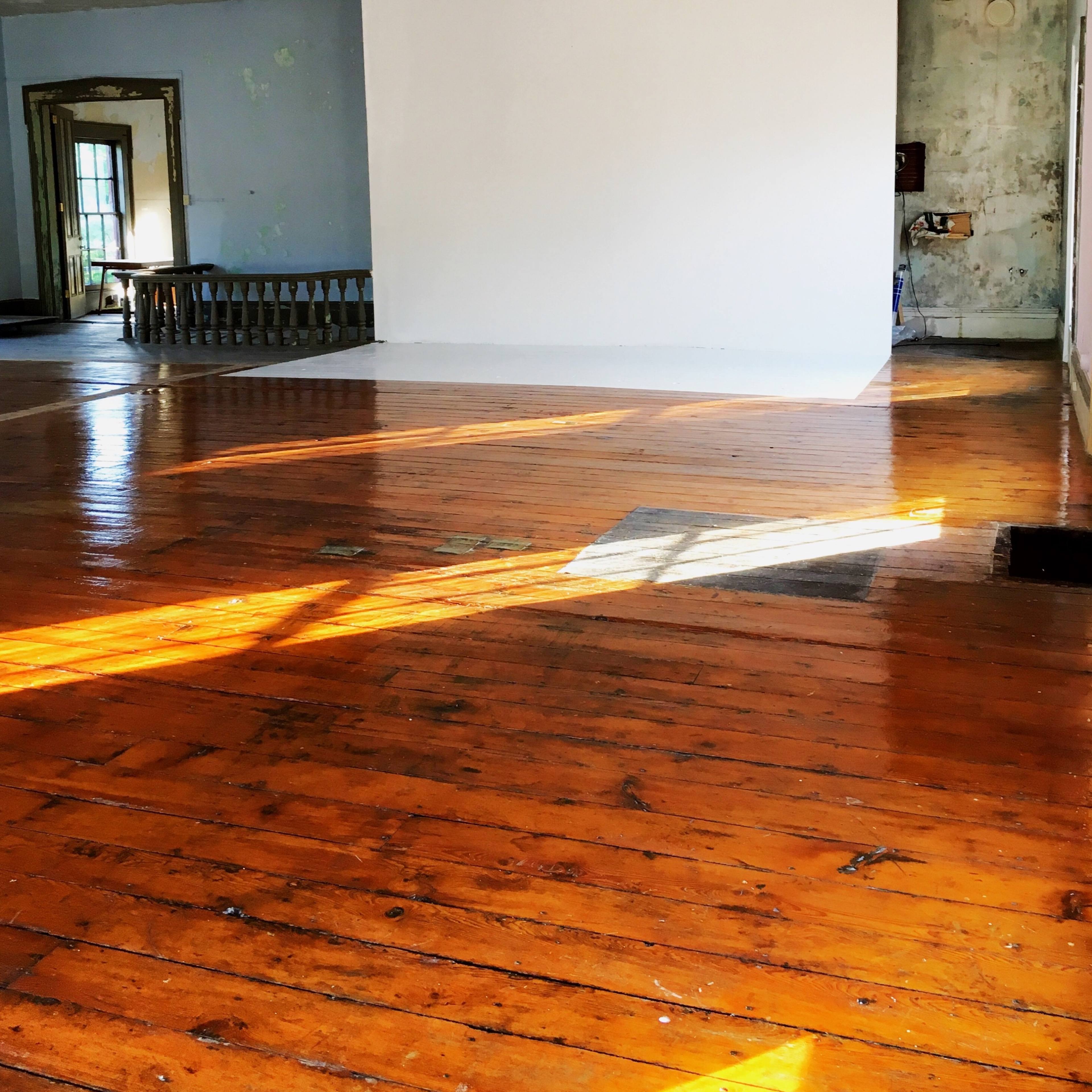 A spacious, empty room with wooden floors, a large white wall, and natural light streaming through the windows.