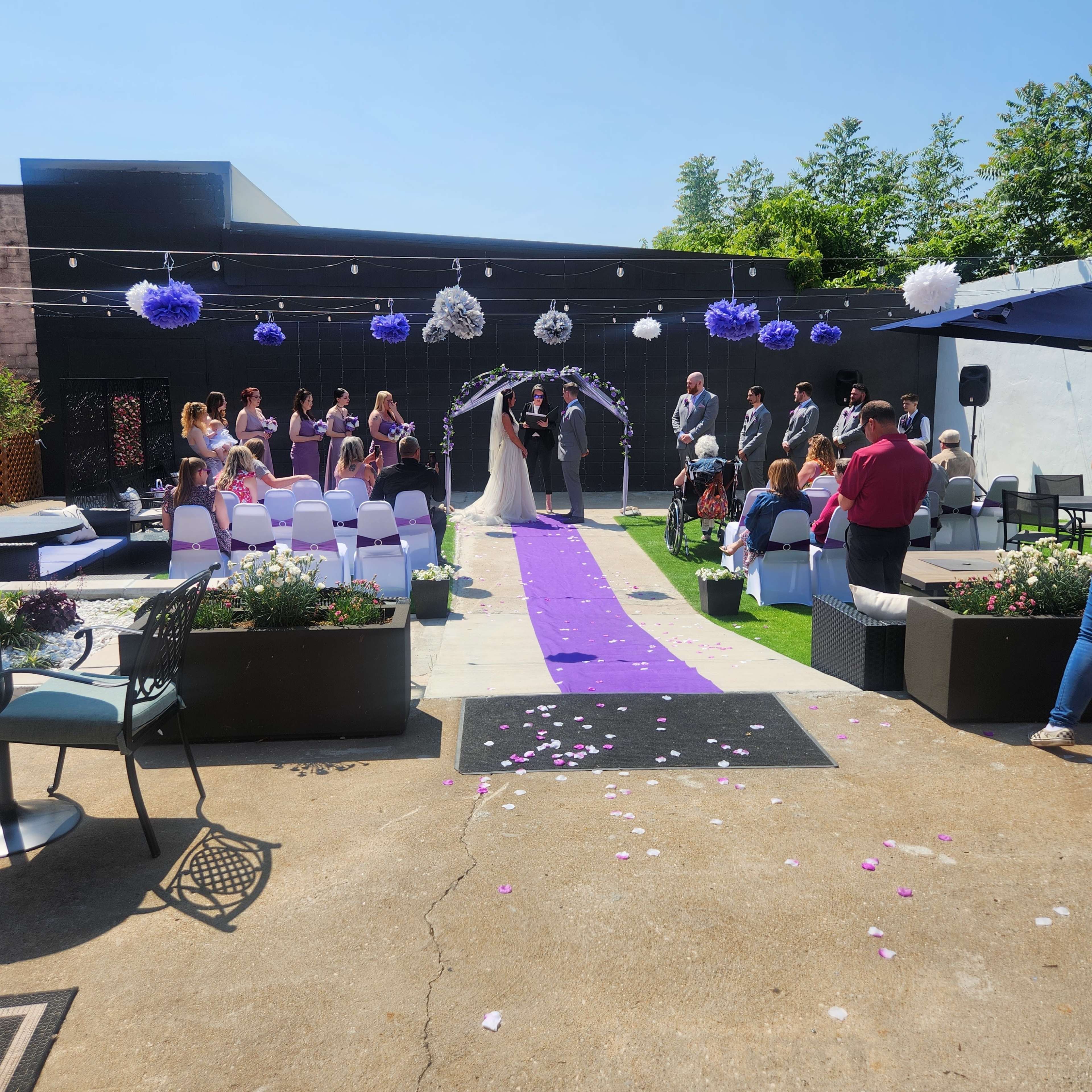 A wedding ceremony is taking place outdoors, with a purple aisle runner, decorated arch, and guests seated on white chairs.