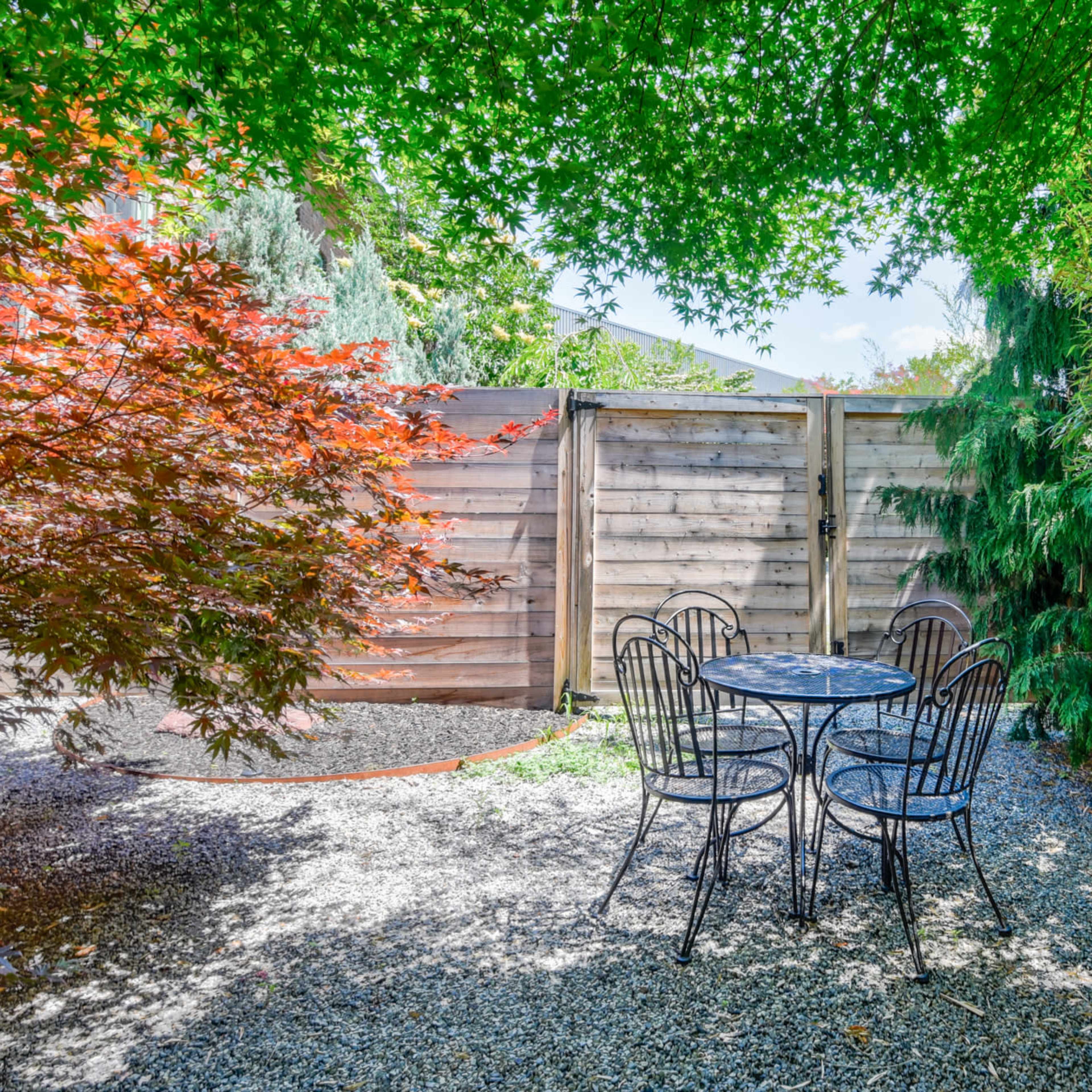 A small outdoor seating area features a black metal table and chairs surrounded by lush greenery and a wooden fence.
