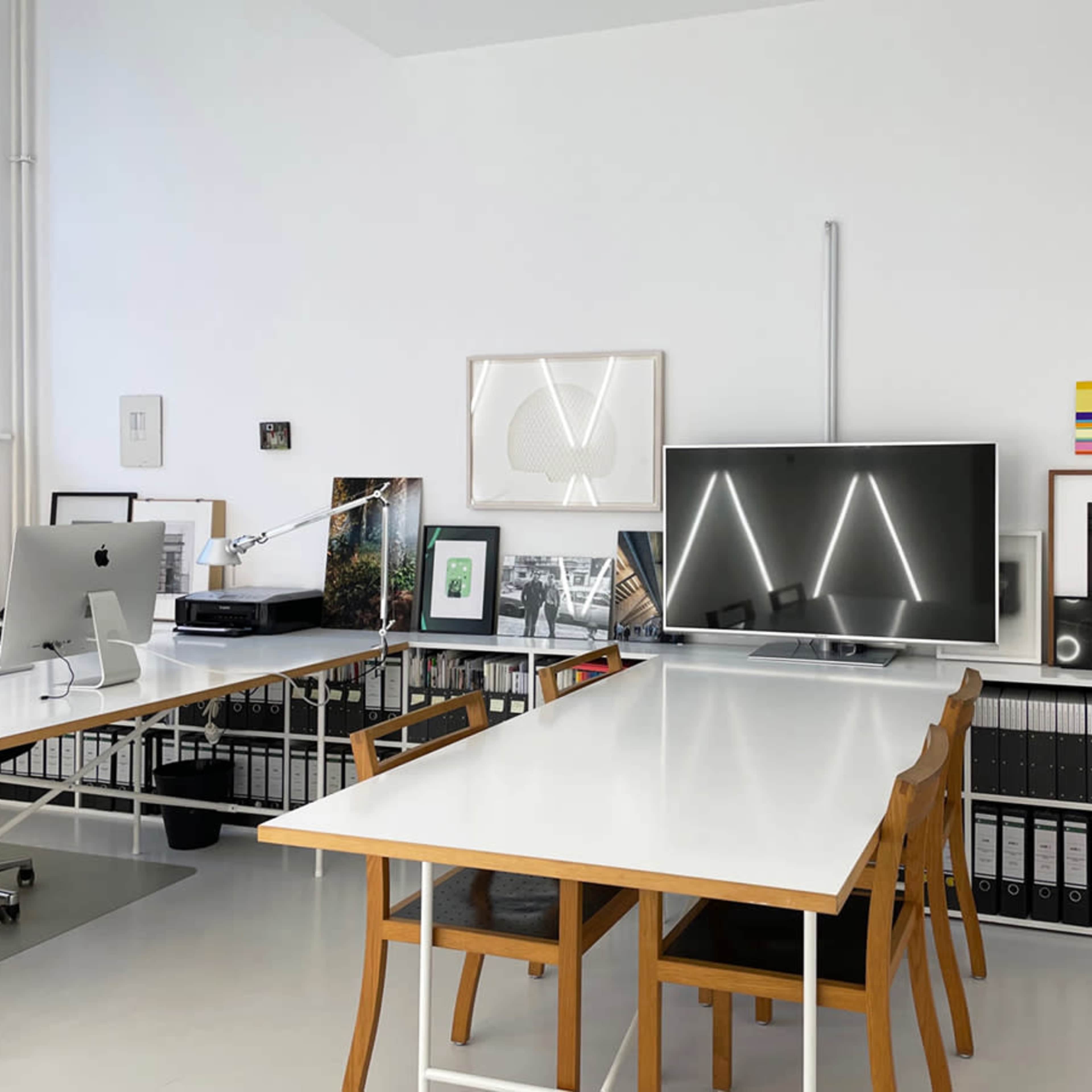 A modern office space features a white interior with a long table, wooden chairs, and organized shelves filled with binders, framed photographs, and a television display.